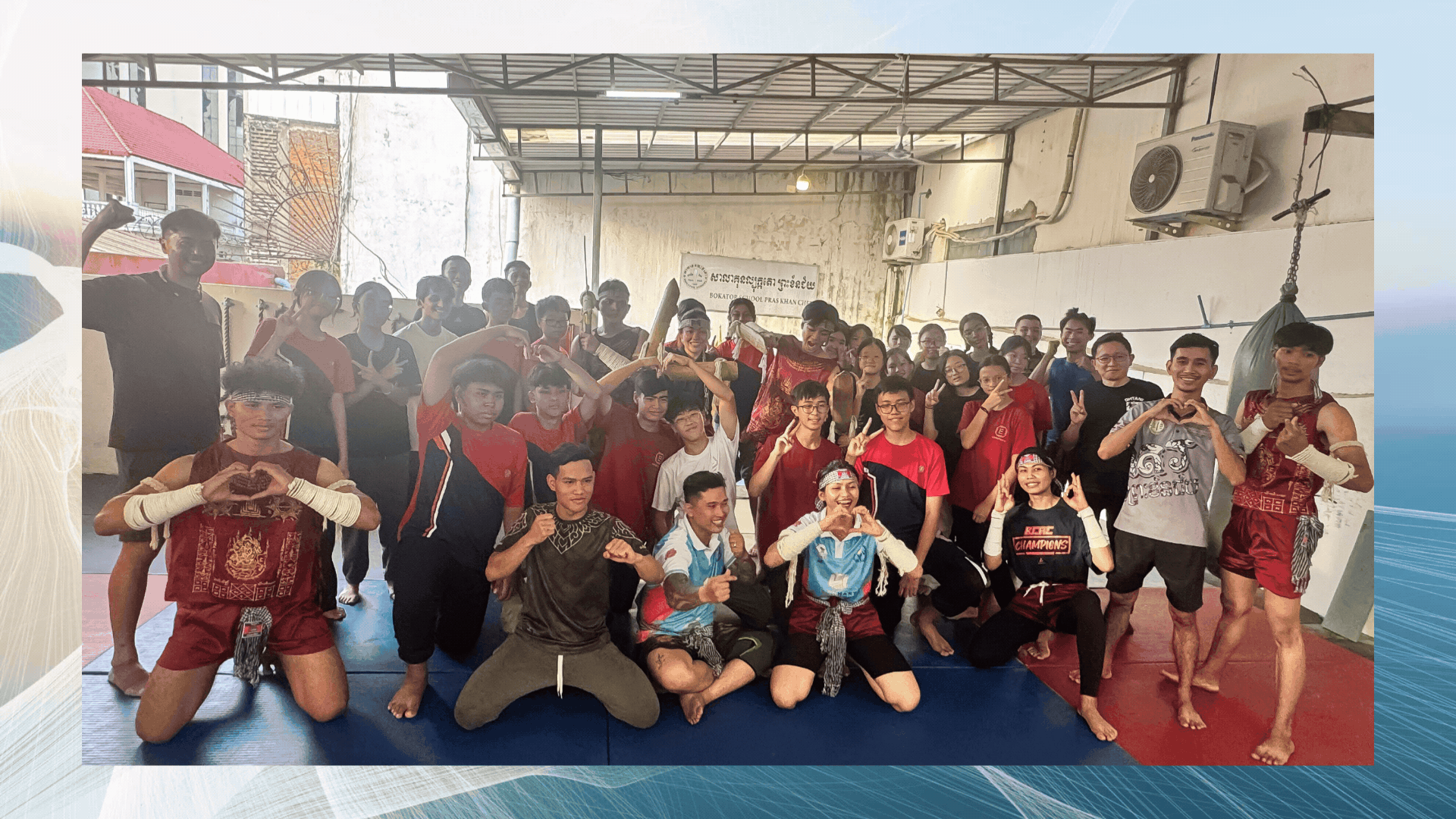 A large group of martial arts students and fighters pose together in a gym, some wearing traditional Khmer boxing attire.