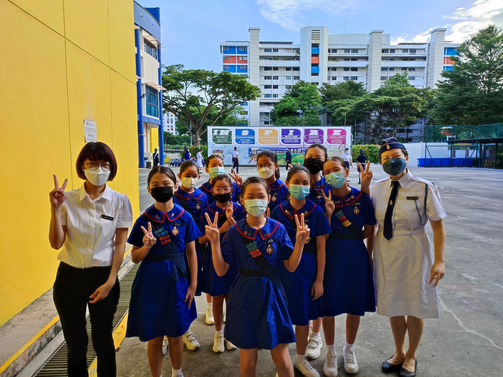 A group of girls in blue uniforms and face masks pose with peace signs in a school courtyard.