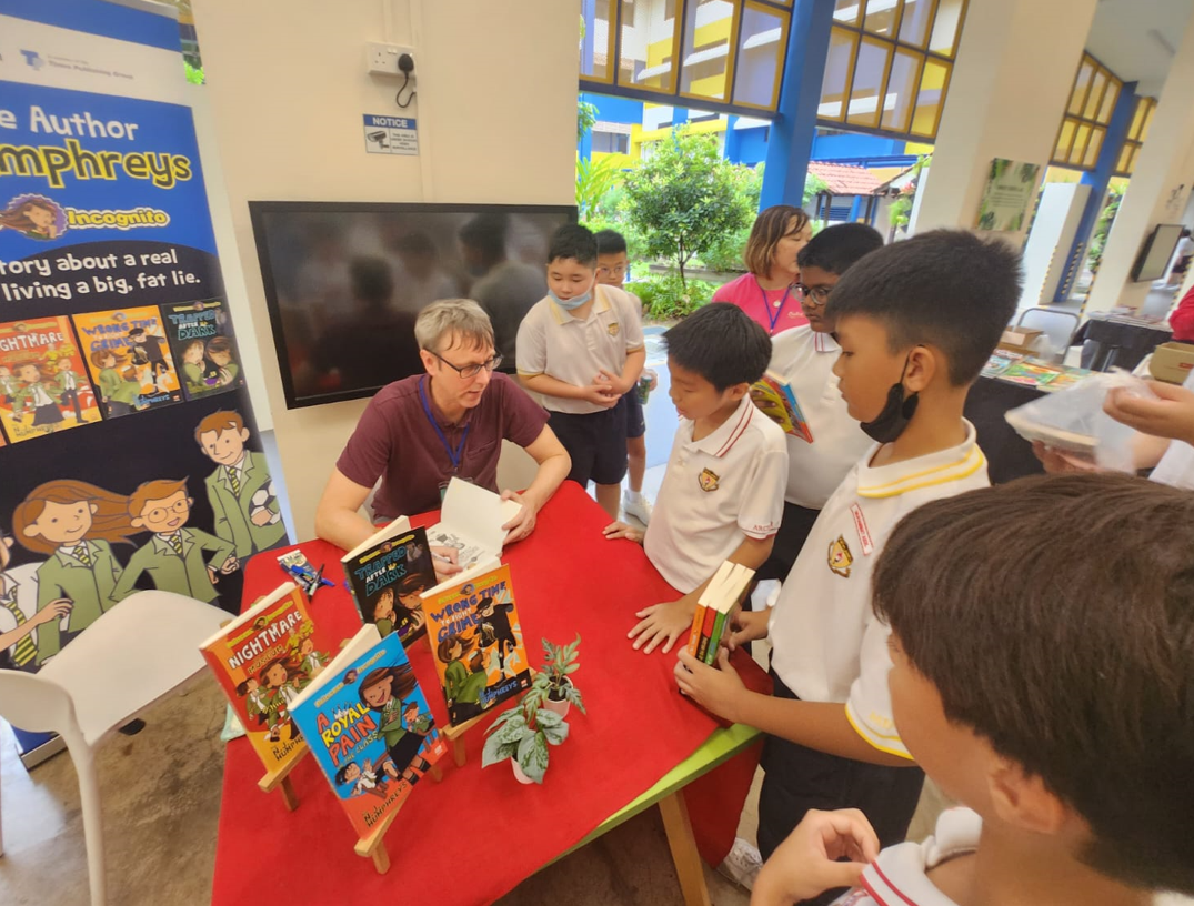 An adult author signs books at a red table surrounded by school children, with a promotional banner nearby.