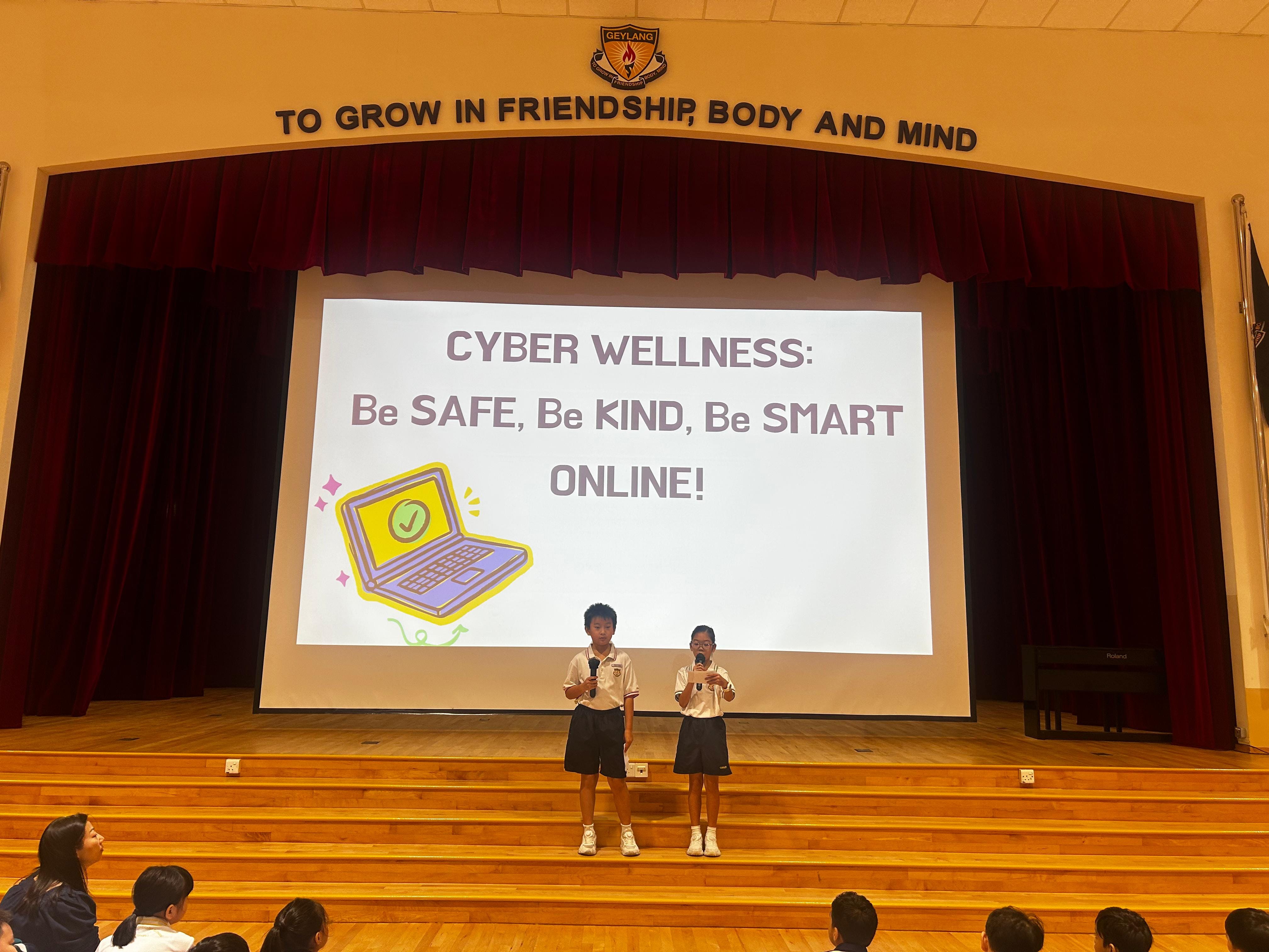 Two student emcees stand on a school hall stage holding microphones, presenting in front of a projected slide that reads "Cyber Wellness: Be Safe, Be Kind, Be Smart Online!" with an illustration of a laptop. The school motto "To Grow in Friendship, Body and Mind" is displayed above the stage.