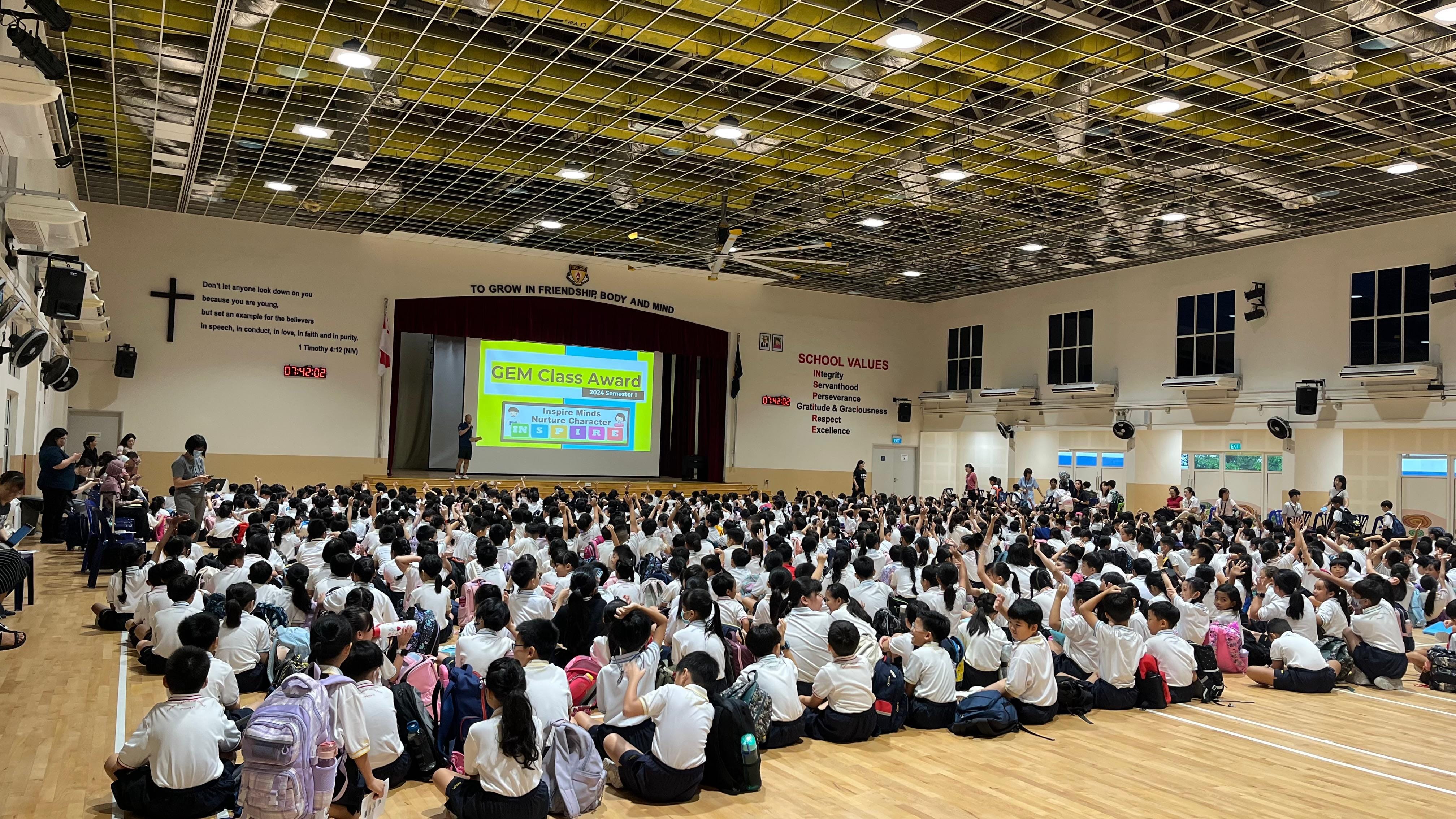 Students in white uniforms sit on a school hall floor facing a screen displaying GEM Class Award presentation.