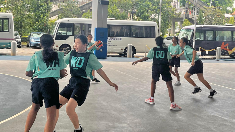 Photo shows a group of young schoolgirls in teal jerseys with position bibs playing a netball match on an outdoor court. One player wearing a "WD" (Wing Defence) bib holds the ball while teammates and opponents in "GA" and "GD" bibs move around her. White minibuses and trees are visible in the background.