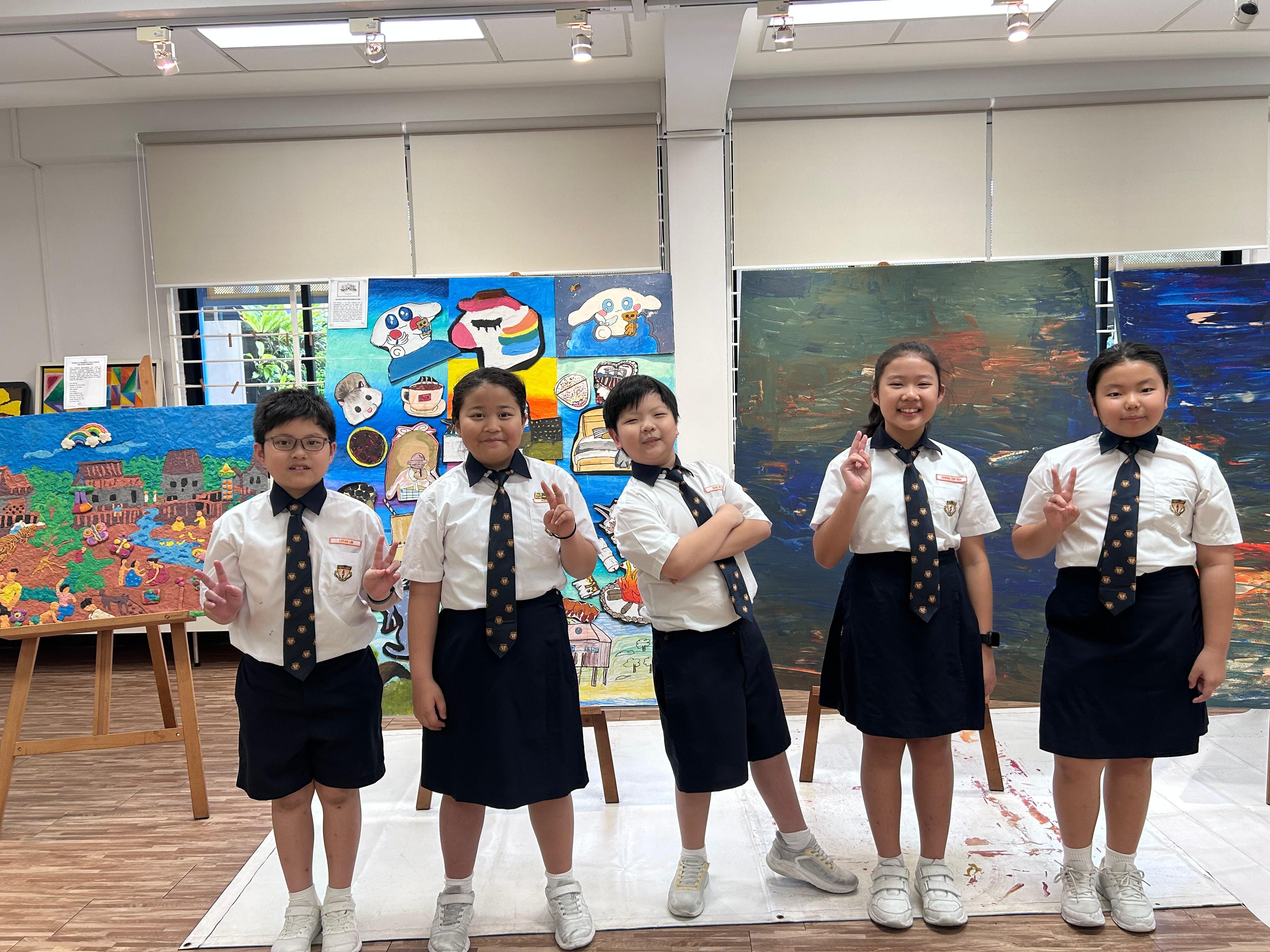 Five uniformed school children pose with peace signs in front of colorful student artwork displayed in an art room.