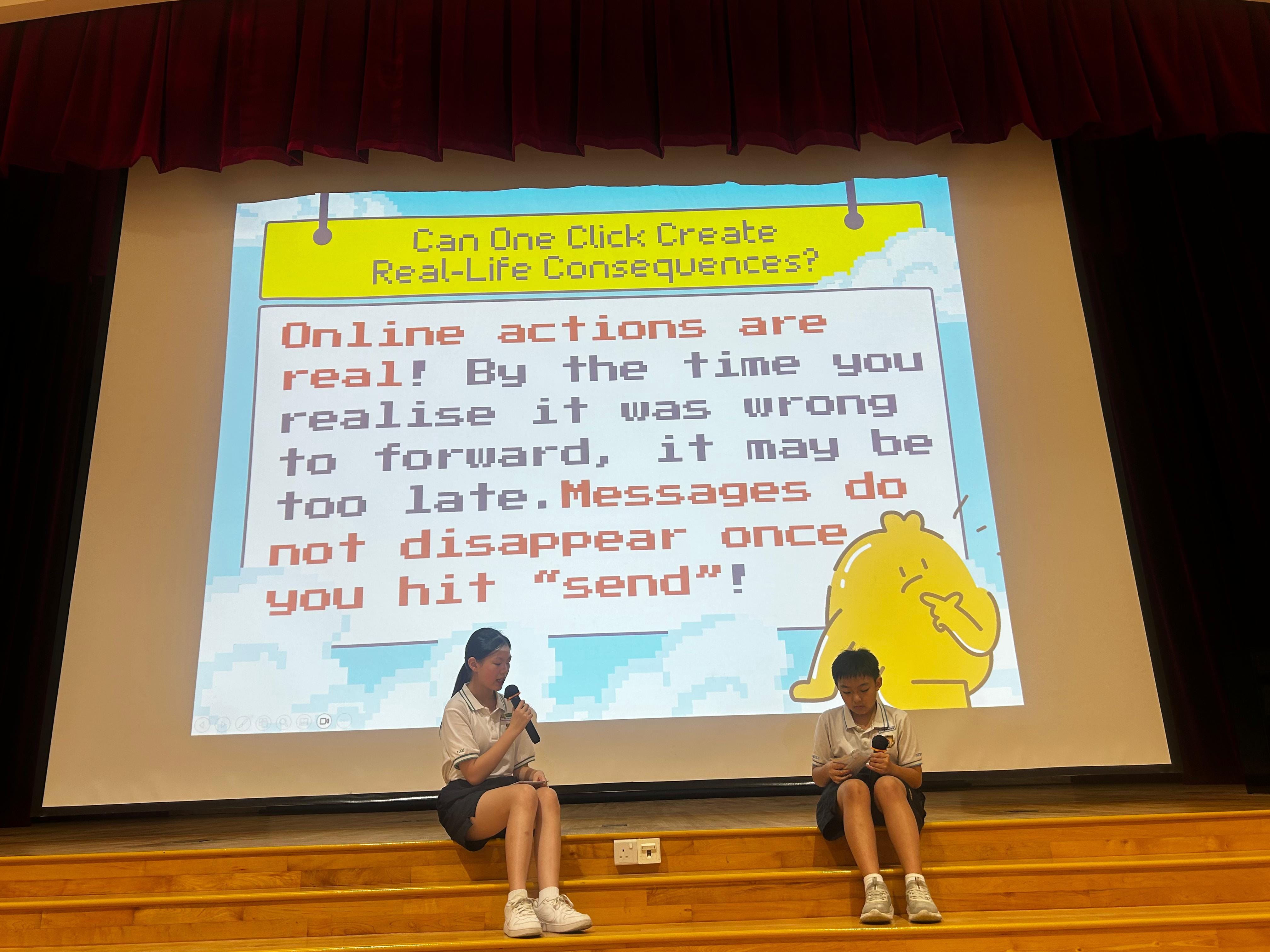 Two student presenters sit on stage in front of a projected slide titled "Can One Click Create Real-Life Consequences?", which reminds students that online actions are real and that messages do not disappear once sent.