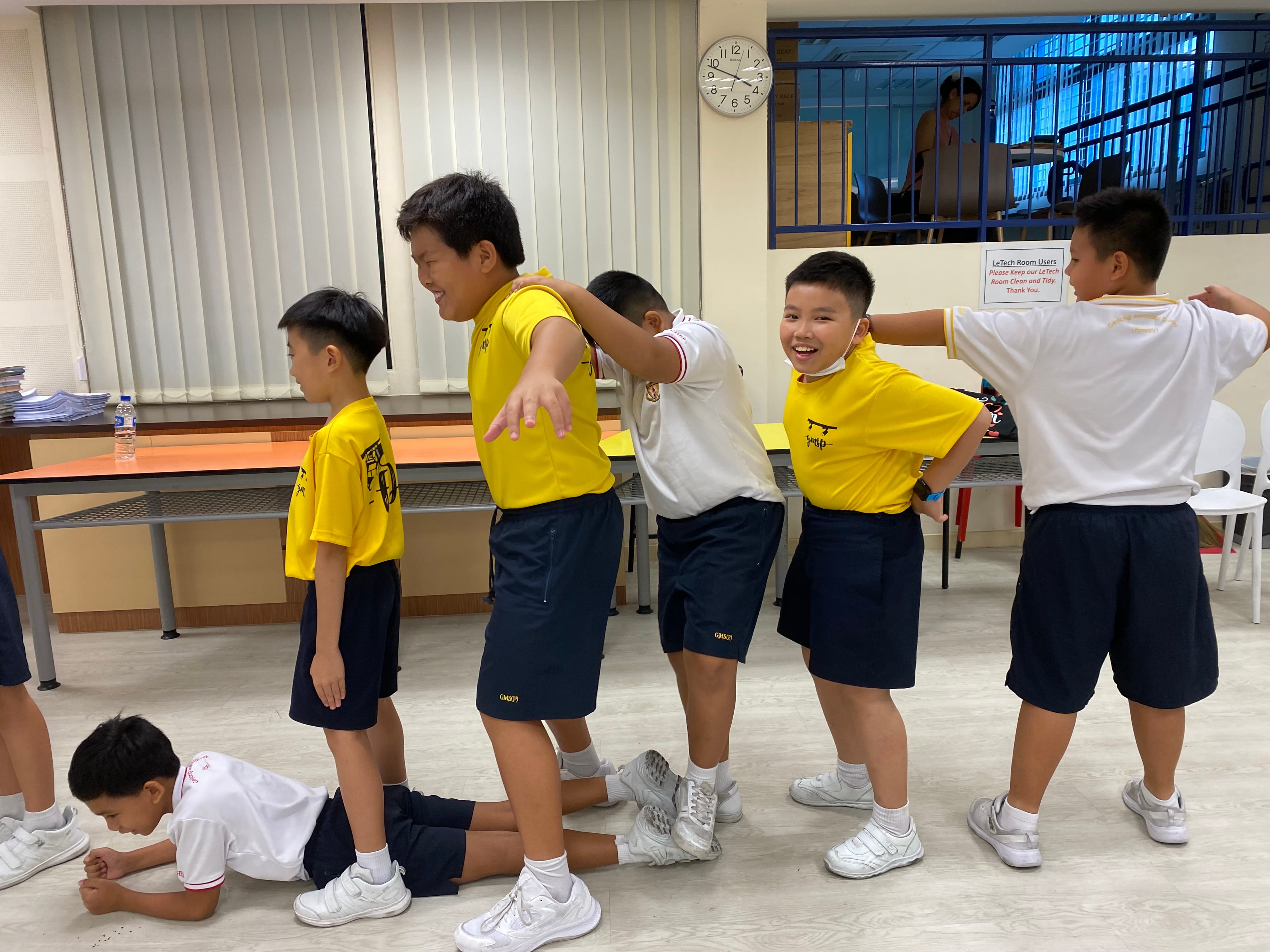Several school boys in yellow and navy uniforms laugh and play together in an indoor classroom.