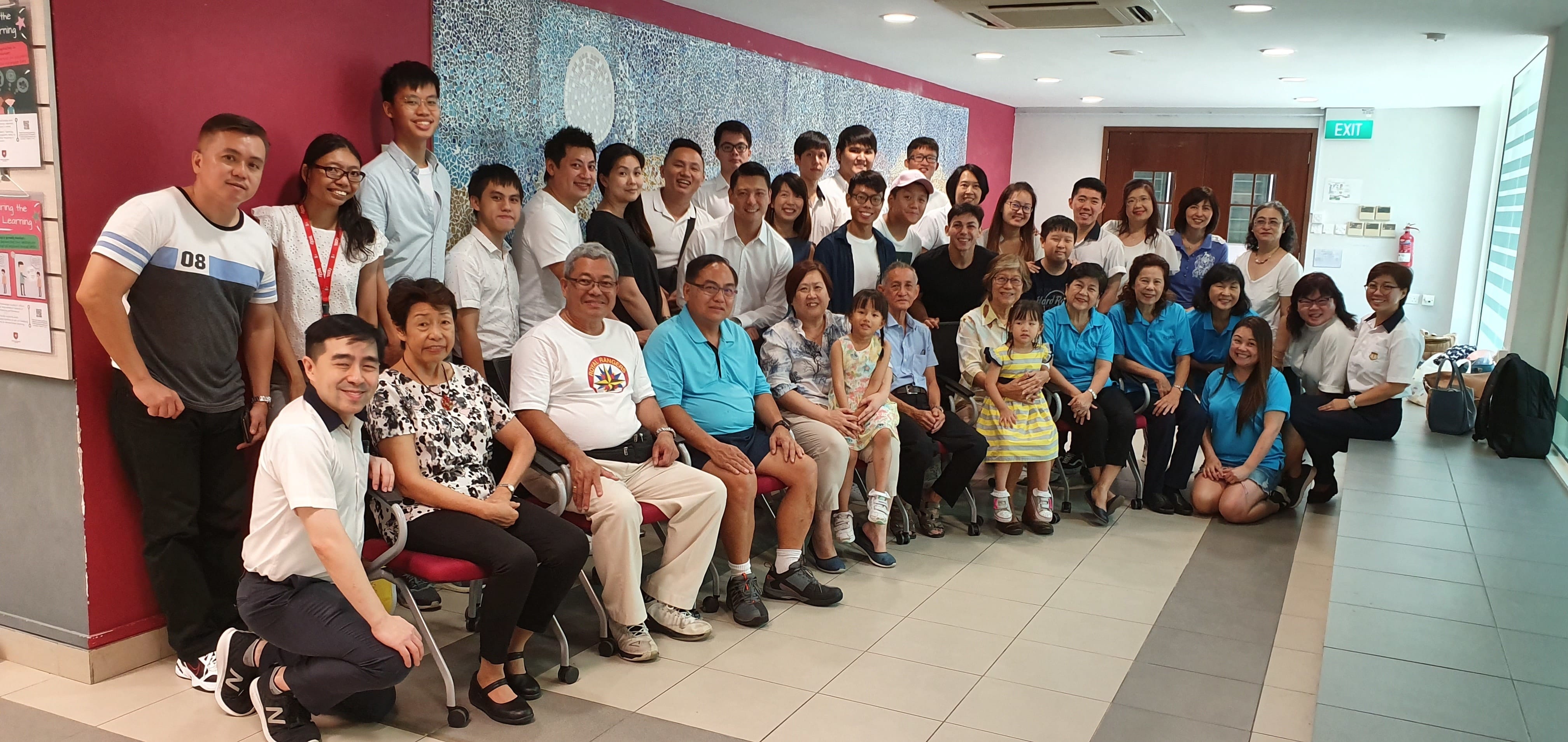 Large group of adults and two young children posing together indoors near a pink wall and decorative mural.