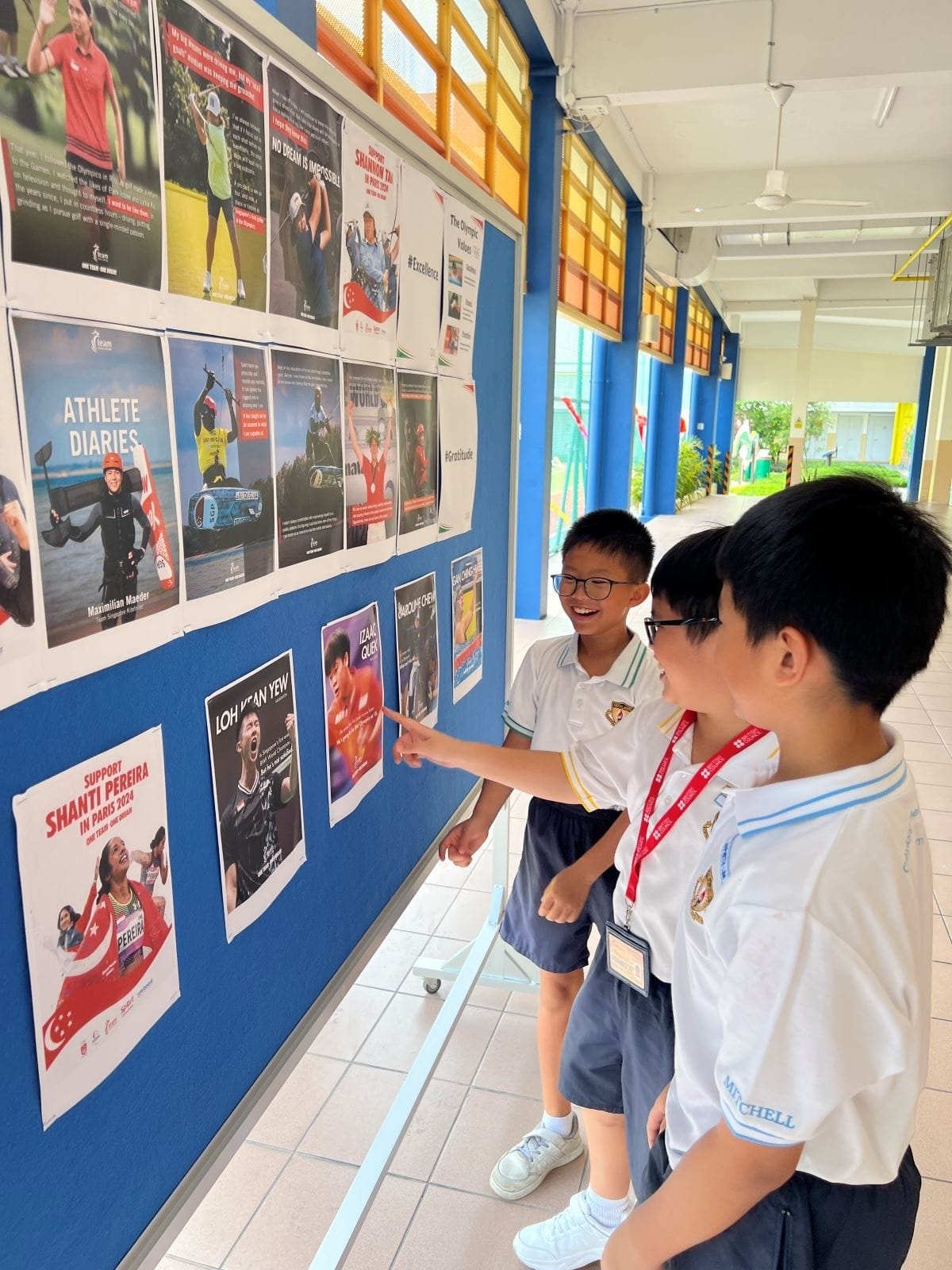 Two primary school students in white uniforms lean over a display board covered with printed posters and articles about Singapore athletes, including Loh Kean Yew and Shanti Pereira. One student points at a poster while the other looks on with interest.