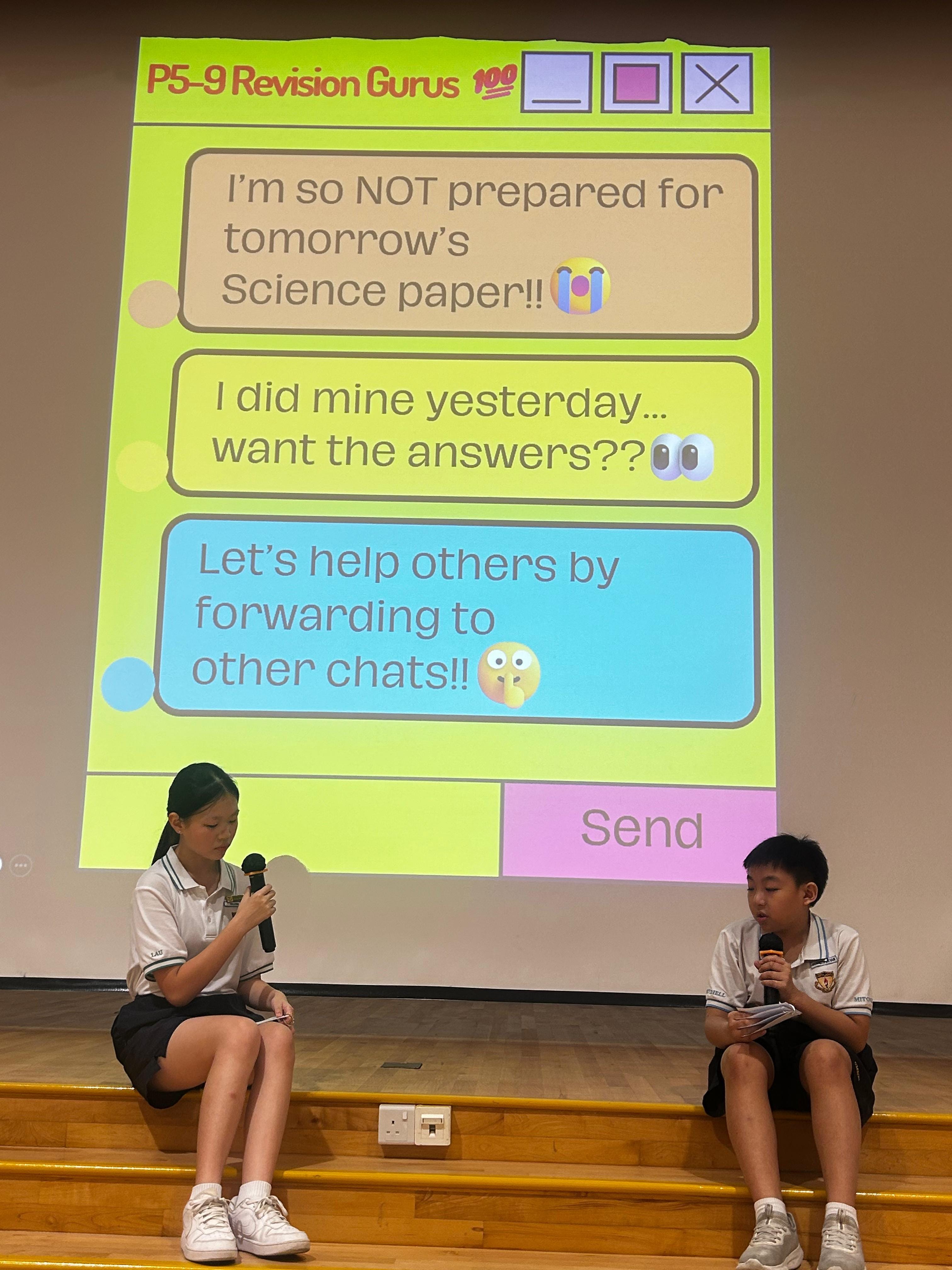 Photo04: Two student performers sit on the edge of a school stage, acting out a skit with a projected chat conversation behind them. The slide, titled "P5-9 Revision Gurus", depicts students discussing sharing exam answers online, illustrating the risks of academic dishonesty in digital spaces.