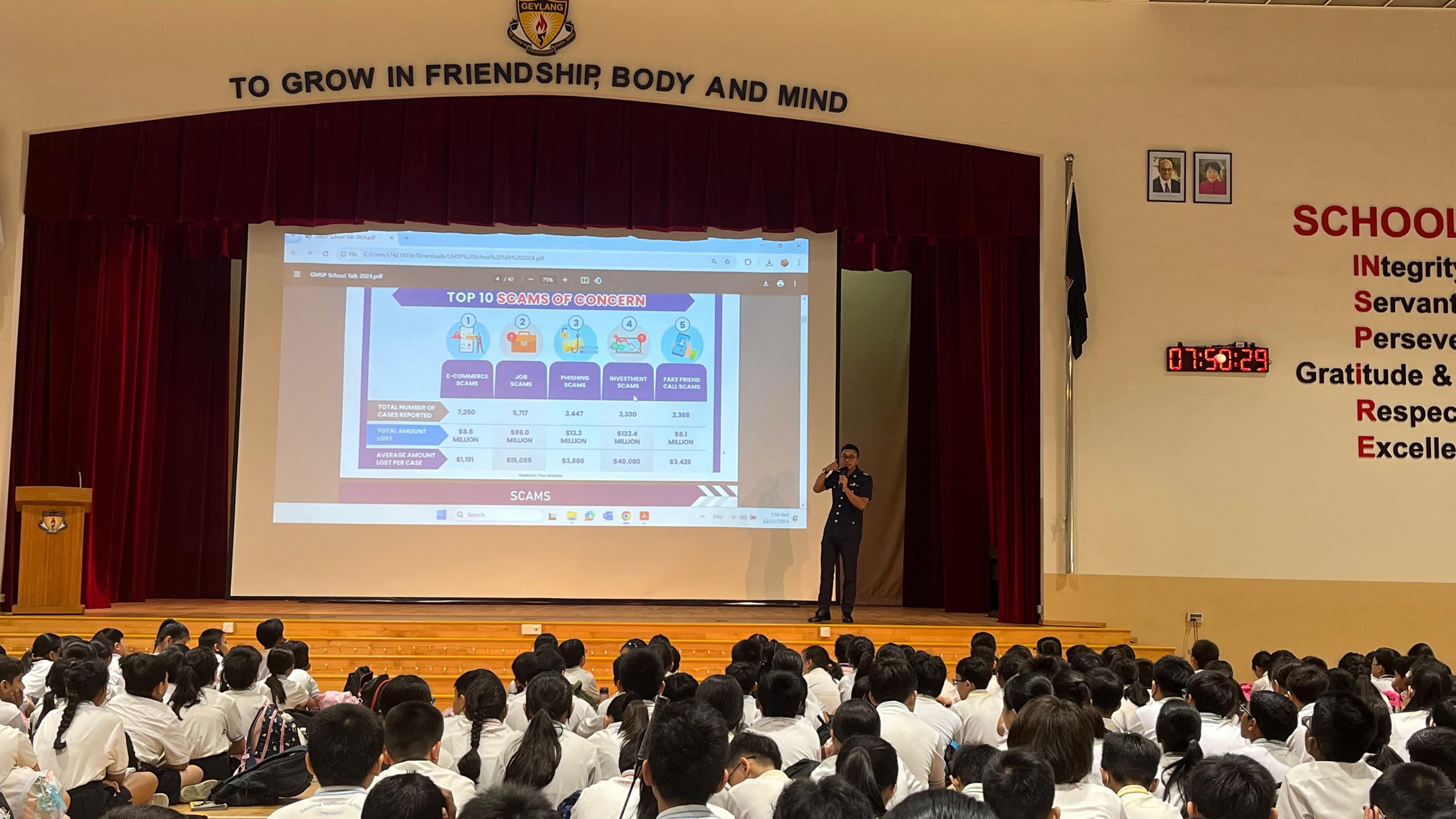 A speaker presents a "Top 10 Scams of Concern" slide to a large school audience in a hall.