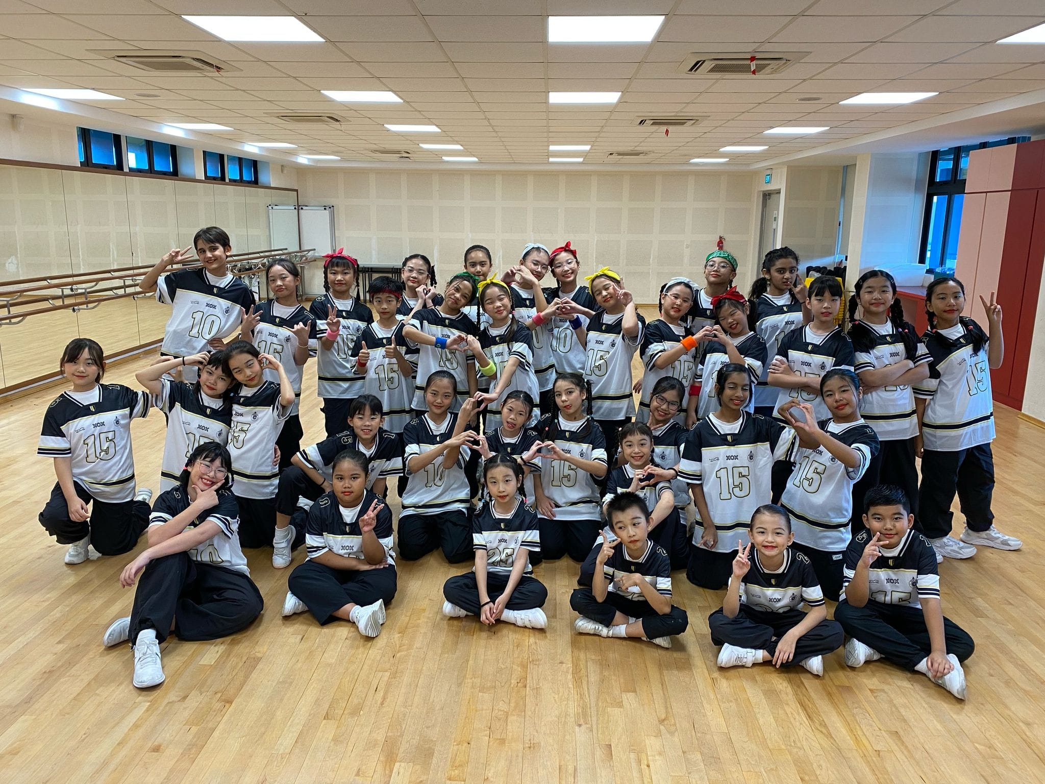 A large group of children in matching black and white numbered jerseys pose together in a dance studio.
