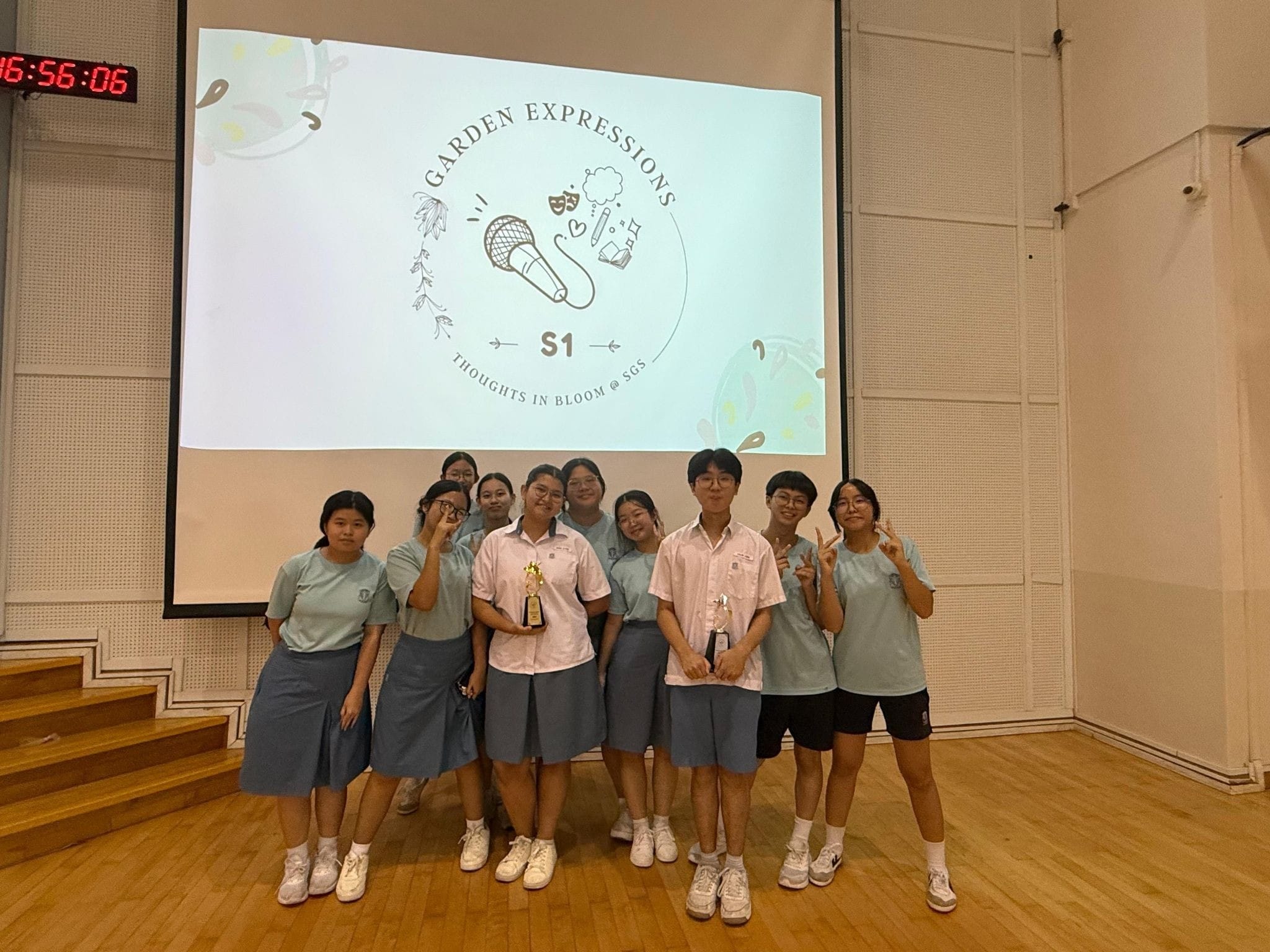 Ten students in school uniforms pose on a stage holding trophies in front of a Garden Expressions S1 projected screen.