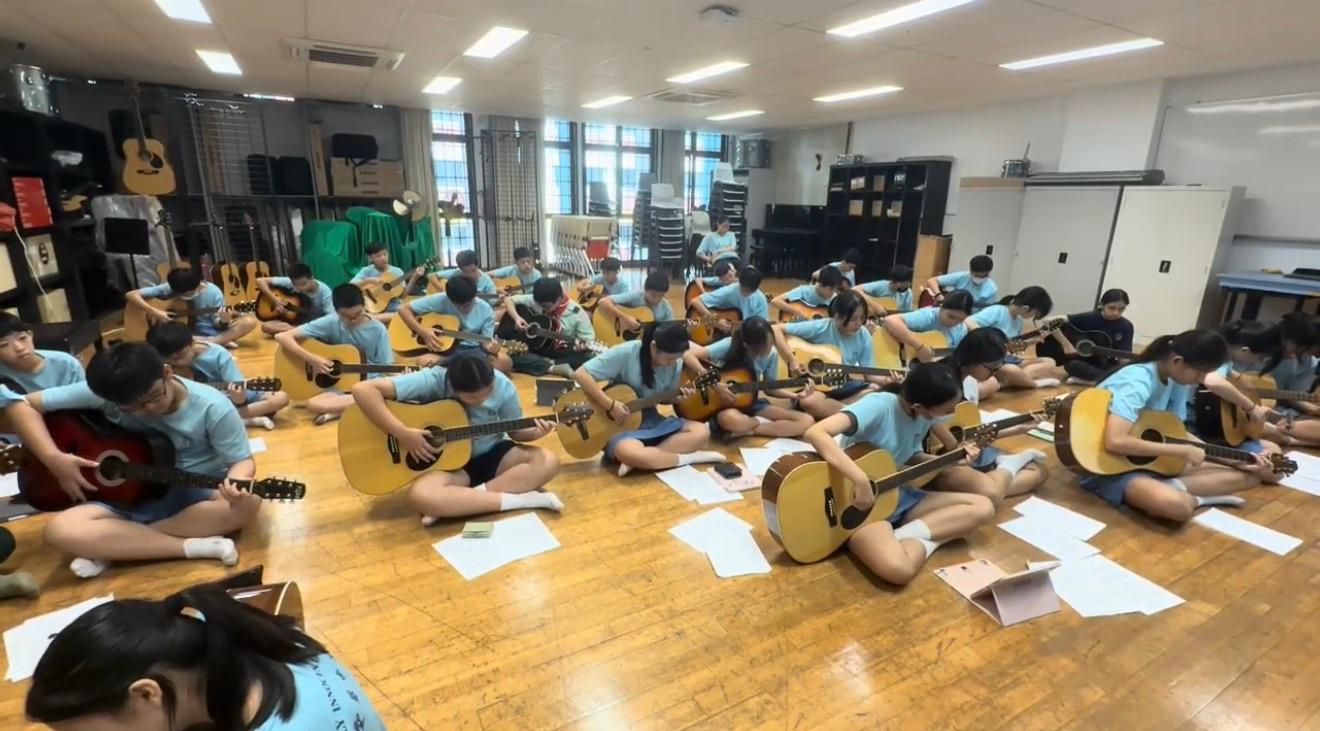Students in blue uniforms sit on a classroom floor playing acoustic guitars with sheet music around them.