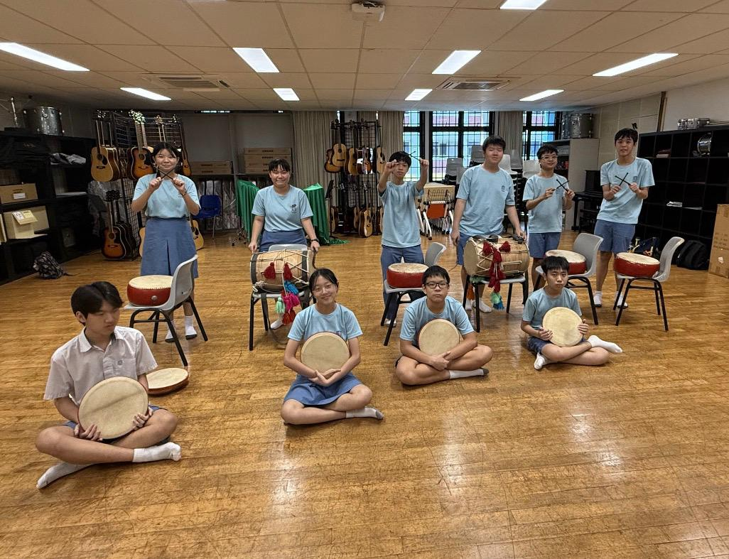 Students in light blue uniforms pose in a music room holding hand drums, with guitars visible on racks behind them.
