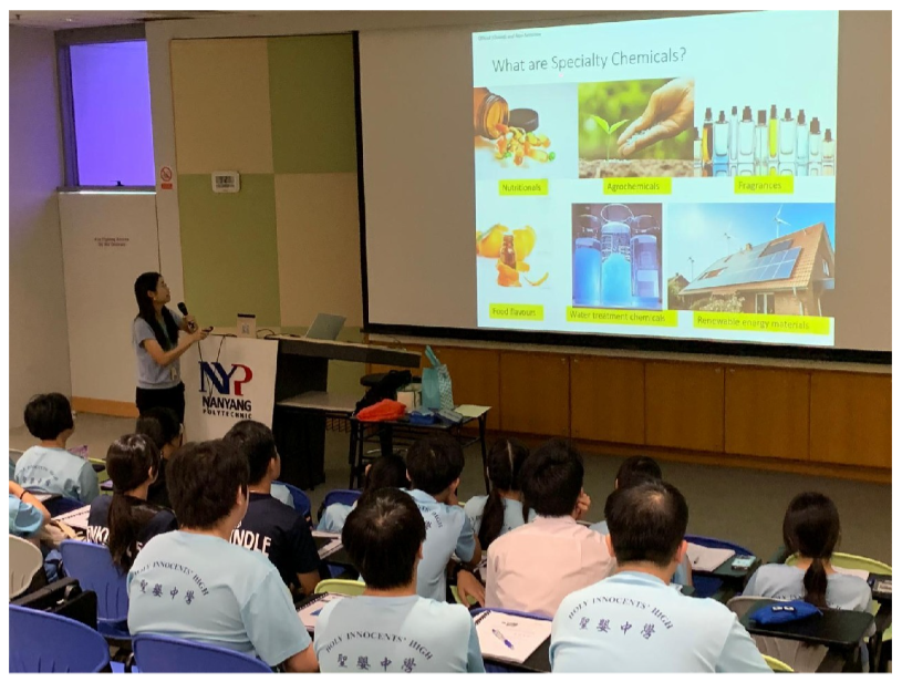 A presenter stands at a Nanyang Polytechnic podium showing a slide titled "What are Specialty Chemicals?" to seated students.