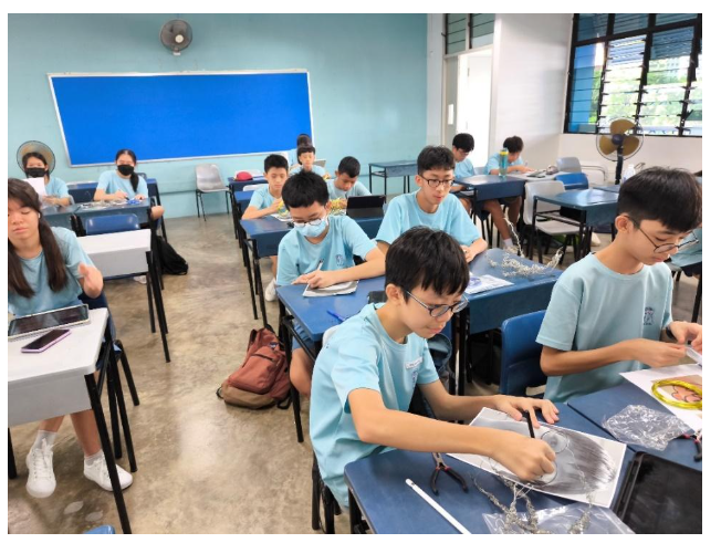 Students in light blue uniforms sit at blue desks in a classroom, working on materials and papers.