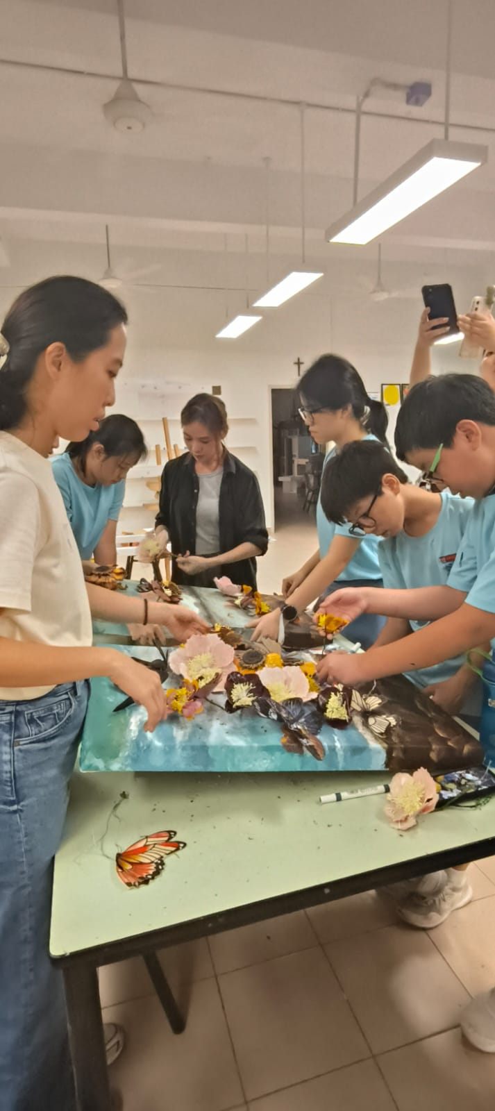 Several students in blue uniforms arrange flowers and butterflies on a blue resin artwork on a table in a classroom.