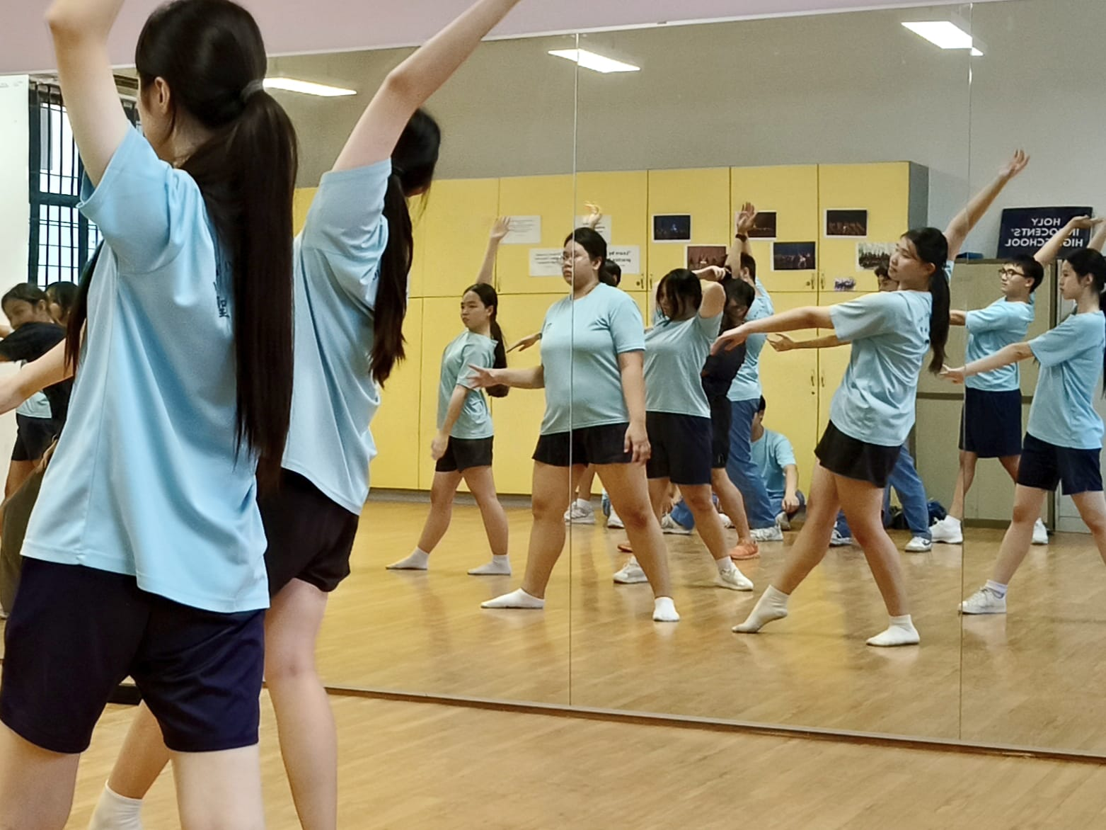 Students in light blue shirts practice dance moves in a studio with large mirrors reflecting the group.