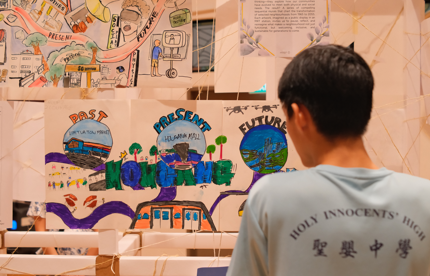 A student in a Holy Innocents' High shirt views a colorful hand-drawn poster about Hougang showing past, present, and future.