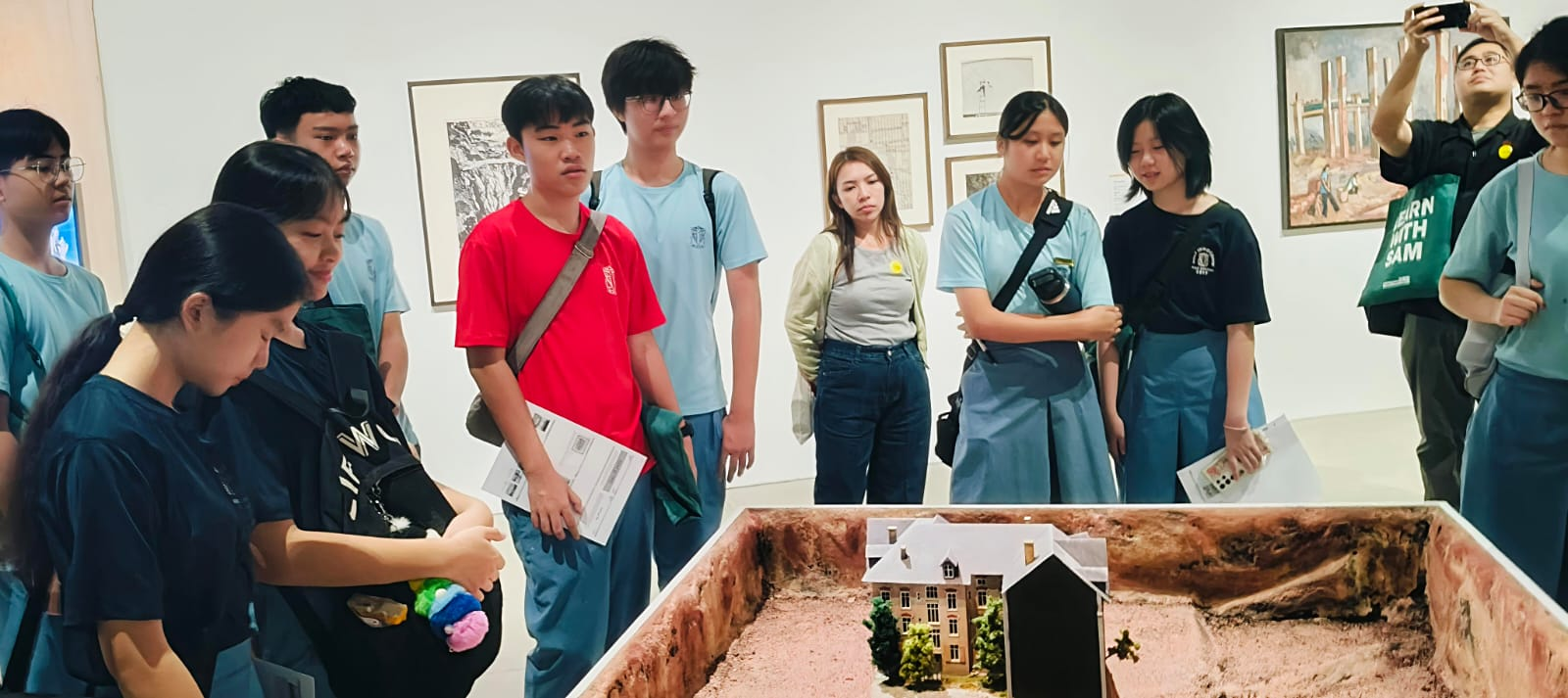 A group of students gathered around a detailed architectural scale model of a building inside an art gallery.