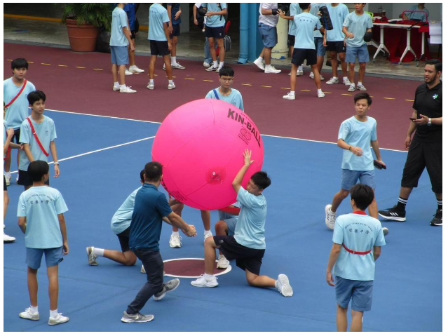 Students in light blue shirts play with a large pink Kin-Ball on an outdoor sports court.