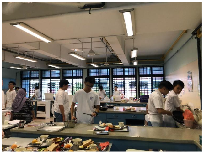 Students in white uniforms cook at counters in a bright school kitchen classroom with large windows.