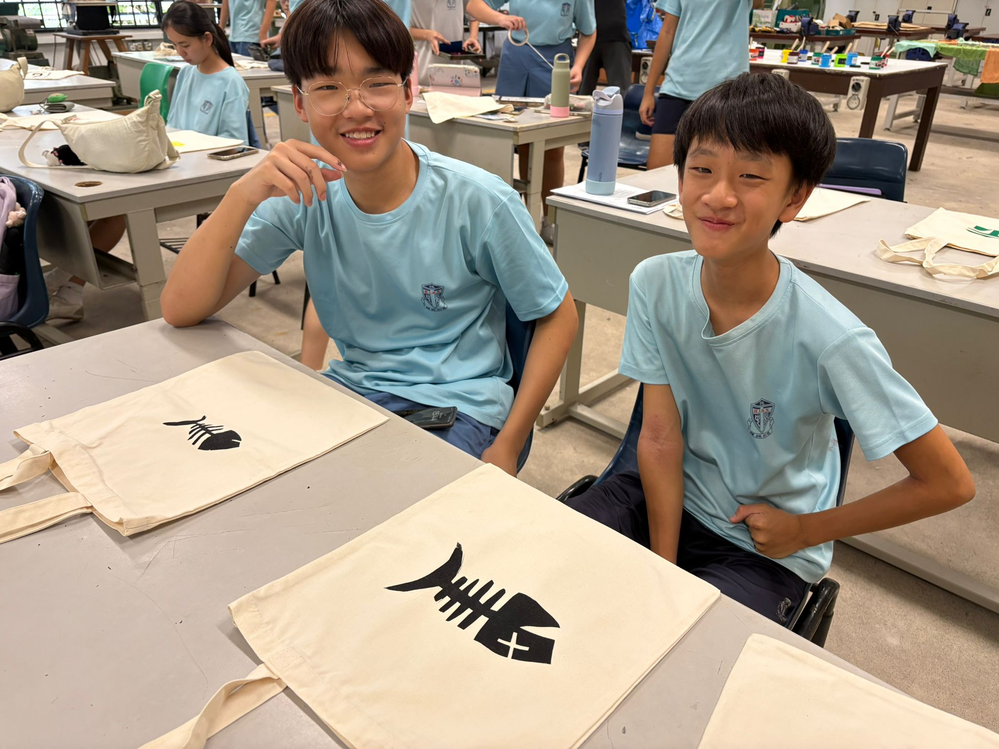 Two smiling students in light blue uniforms sit at a desk with canvas tote bags printed with fish skeleton designs.