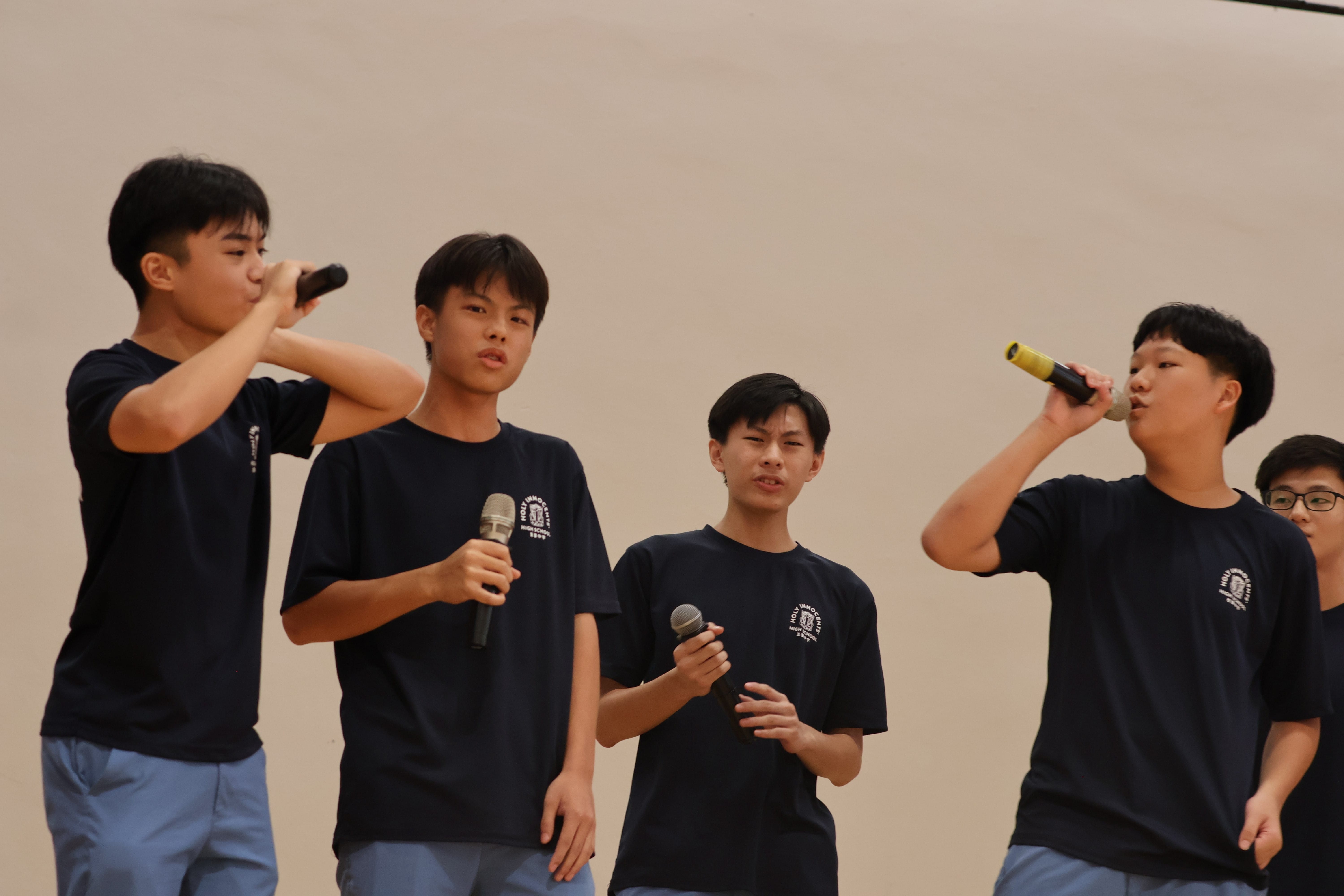 Five teenage boys in matching navy shirts hold microphones and perform on stage together.