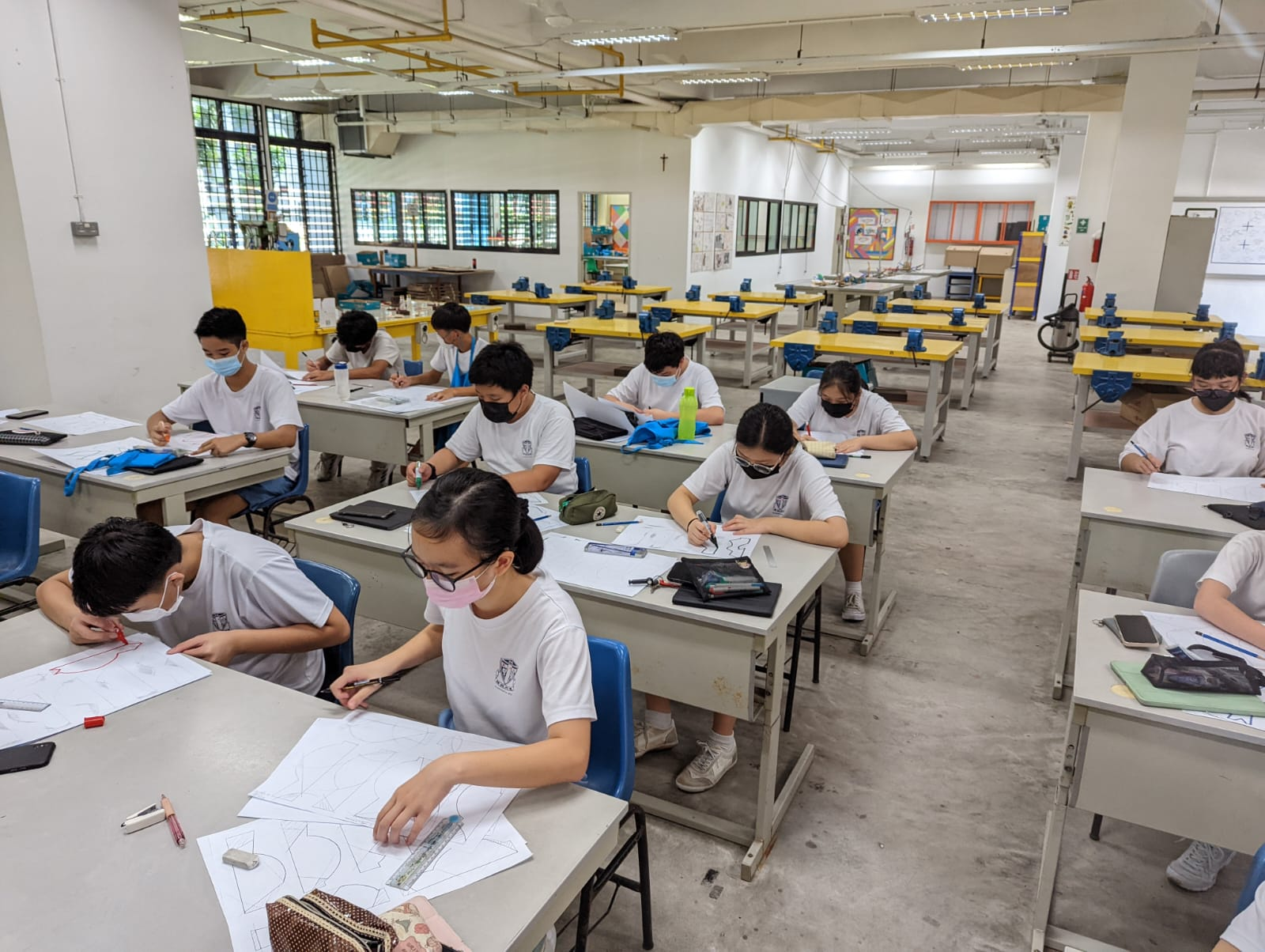 Students in white uniforms and face masks sit at desks in a workshop classroom, drawing geometric shapes on paper.