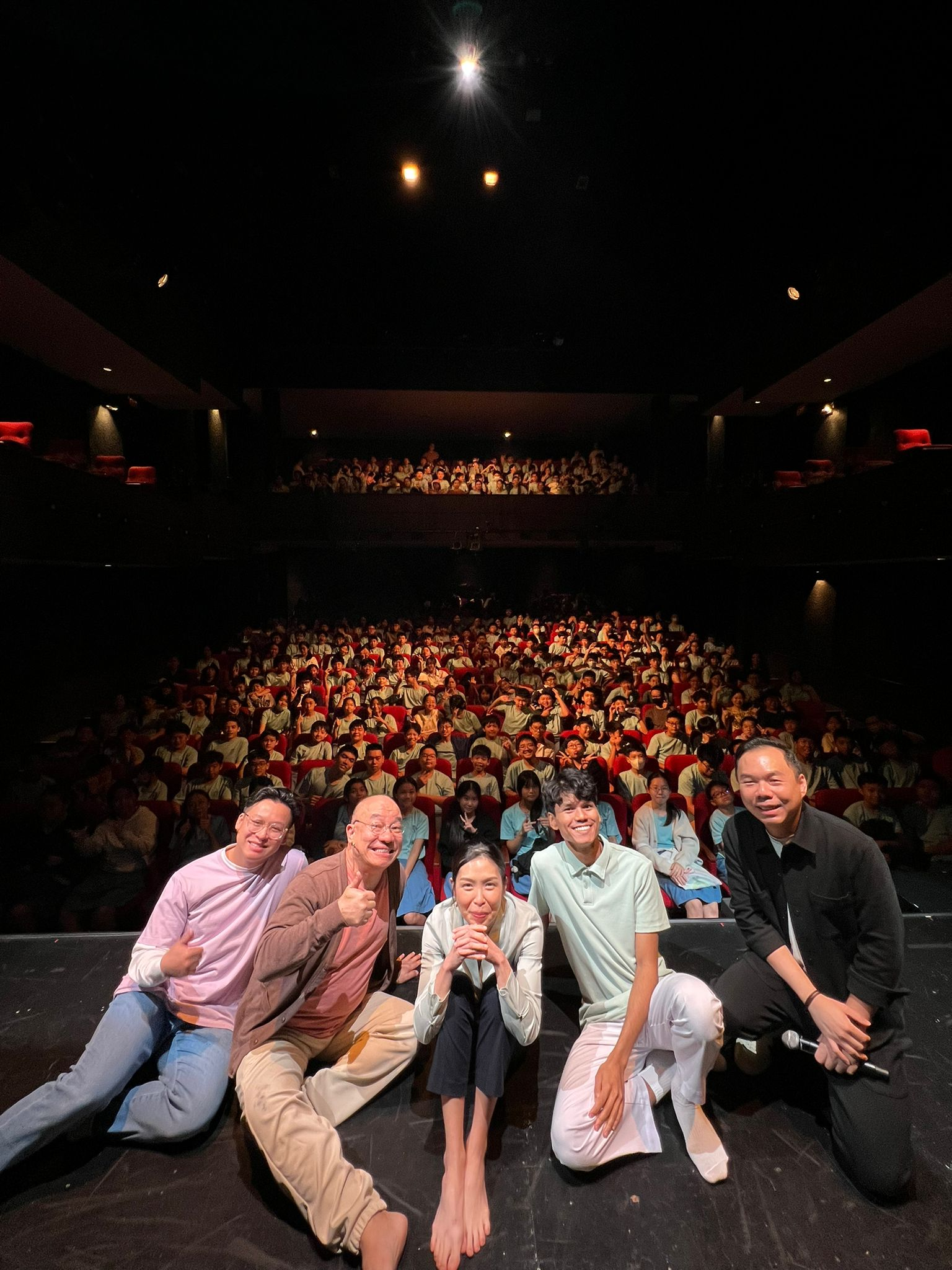 Five people sit and pose on a dark stage in front of a large packed theater audience filling red seats.