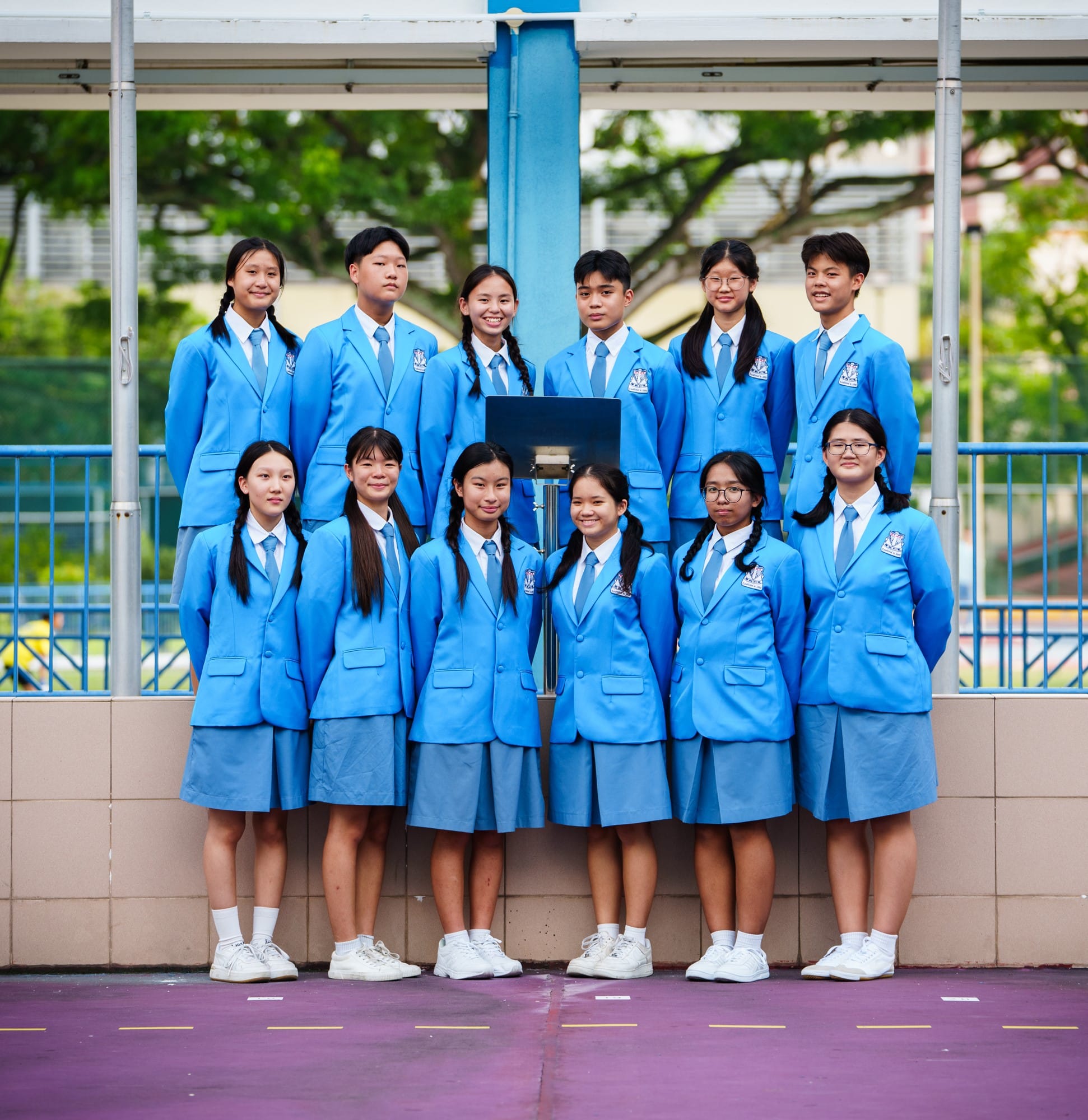 Twelve students in matching blue blazers and ties pose in two rows outdoors at a school courtyard.