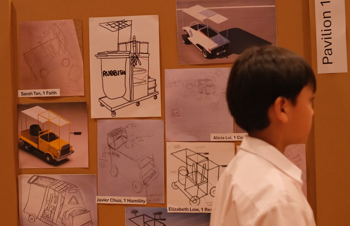 A young boy looks at a display board showing students' vehicle and cart design sketches labeled with their names.
