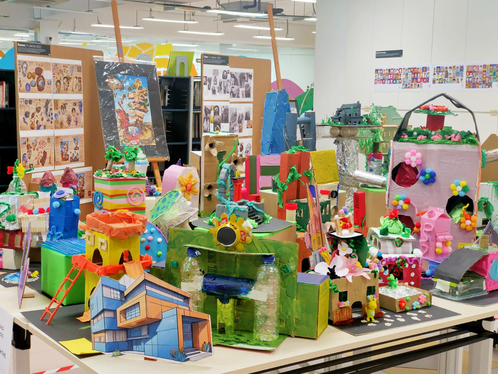 Colorful student art exhibition with cardboard and recycled material 3D model buildings displayed on a table.