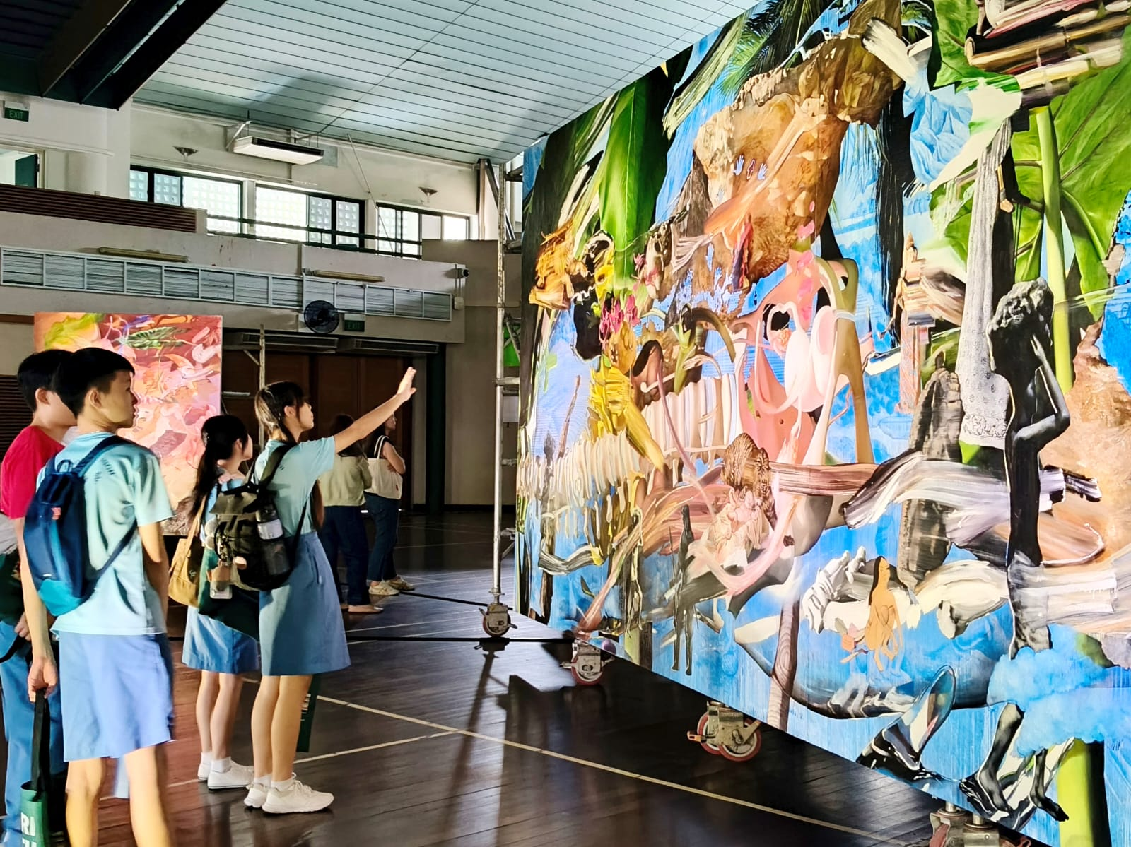 Several visitors stand before a large colorful mural with tropical leaves, figures, and abstract shapes in an indoor hall.