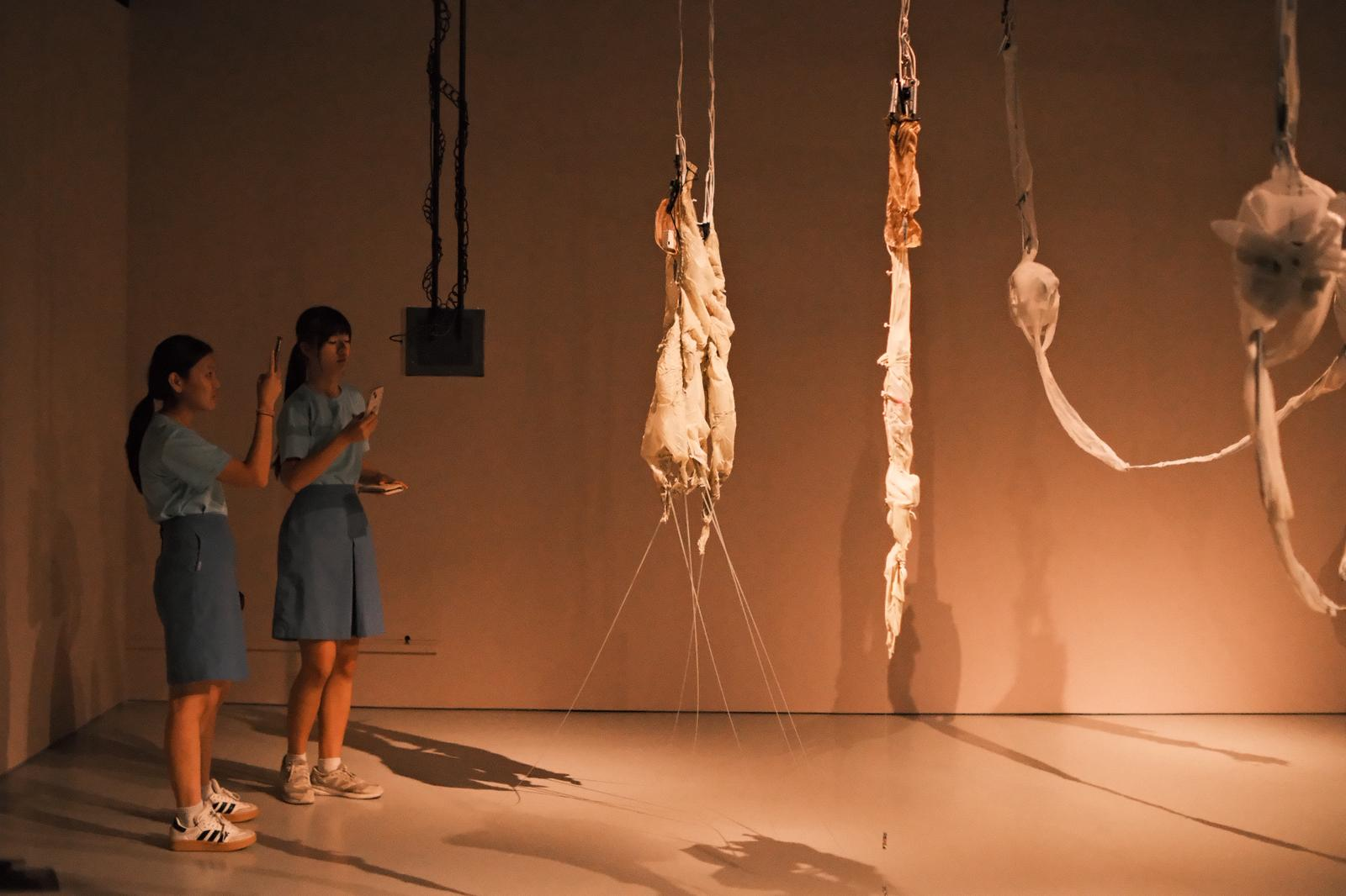 Two students in blue uniforms photograph hanging fabric and rope sculptures in a warmly lit gallery room.