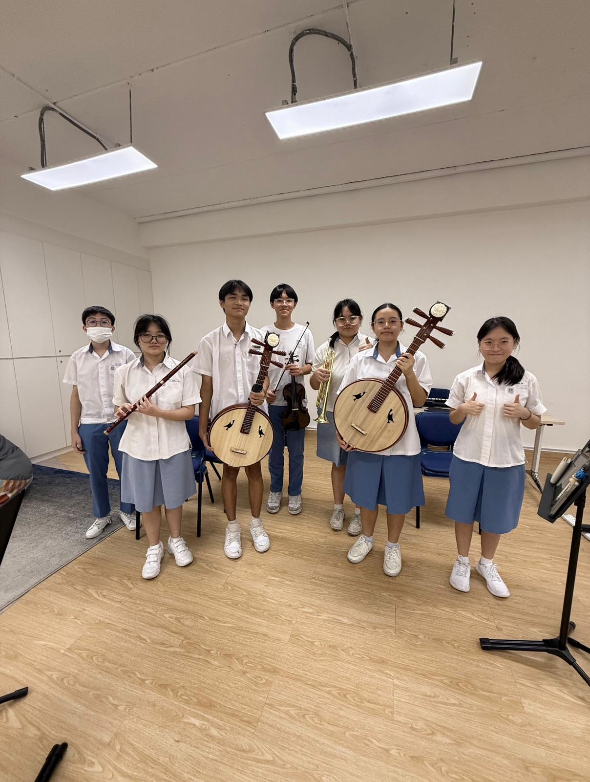 Seven students in white uniforms pose holding traditional Chinese instruments including yueqin, erhu, violin, and flute in a rehearsal room.