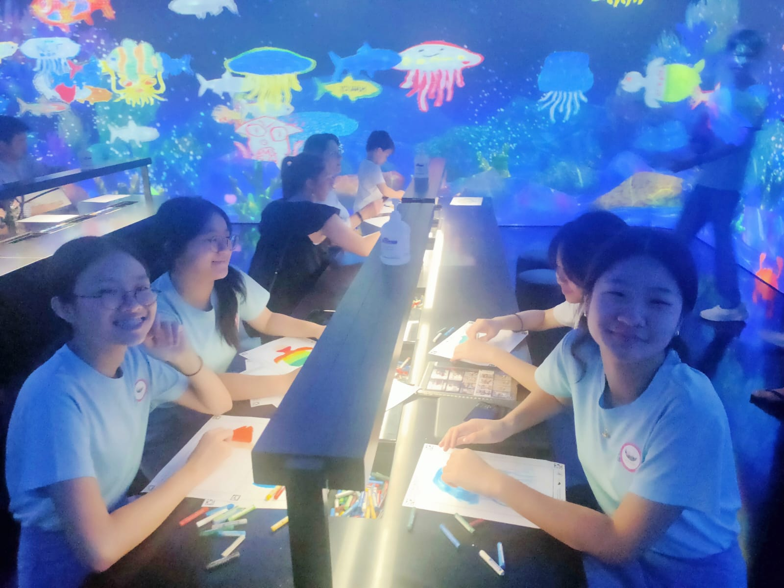Students in blue shirts draw at a lit table surrounded by a colorful projected ocean mural with fish and jellyfish.