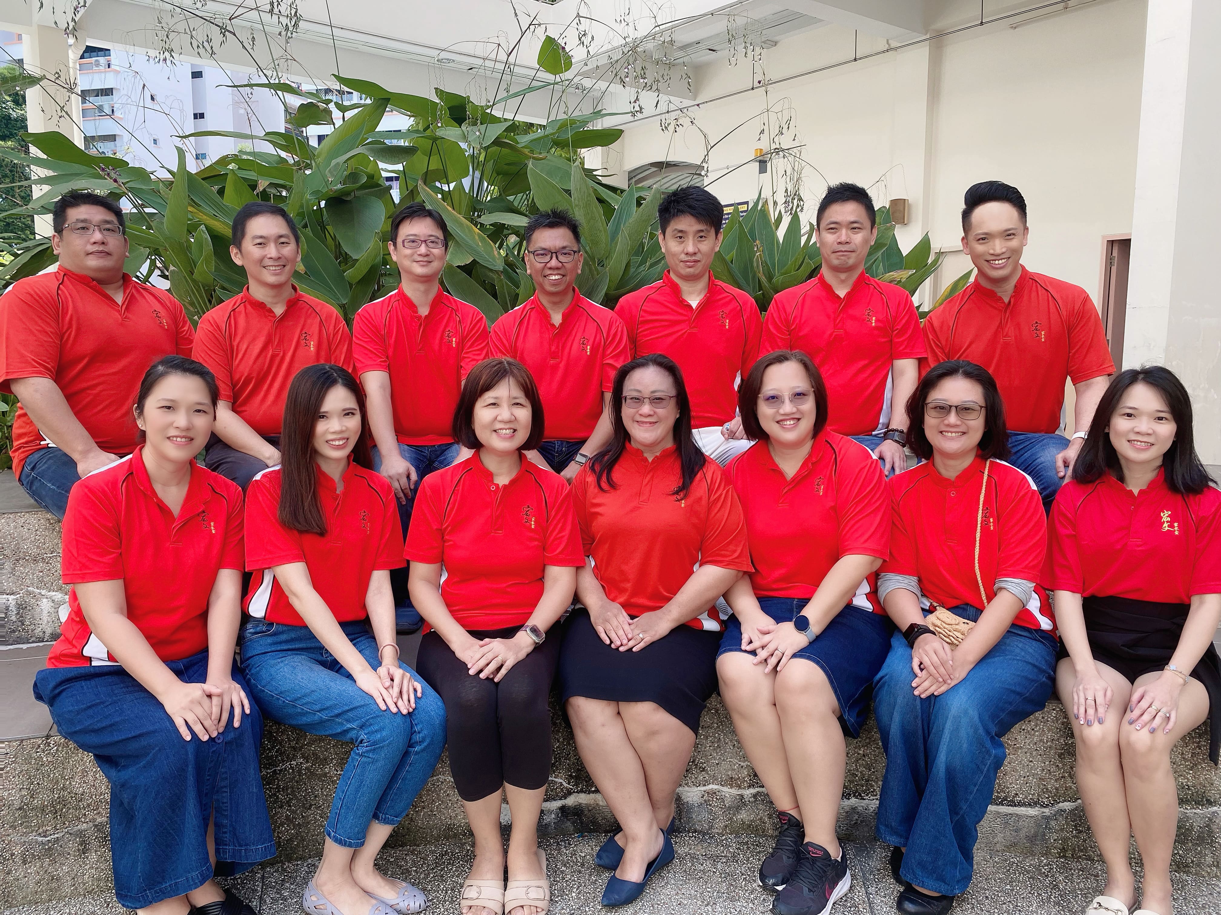 Group of 14 adults in matching red polo shirts posing in two rows outdoors near large green plants.