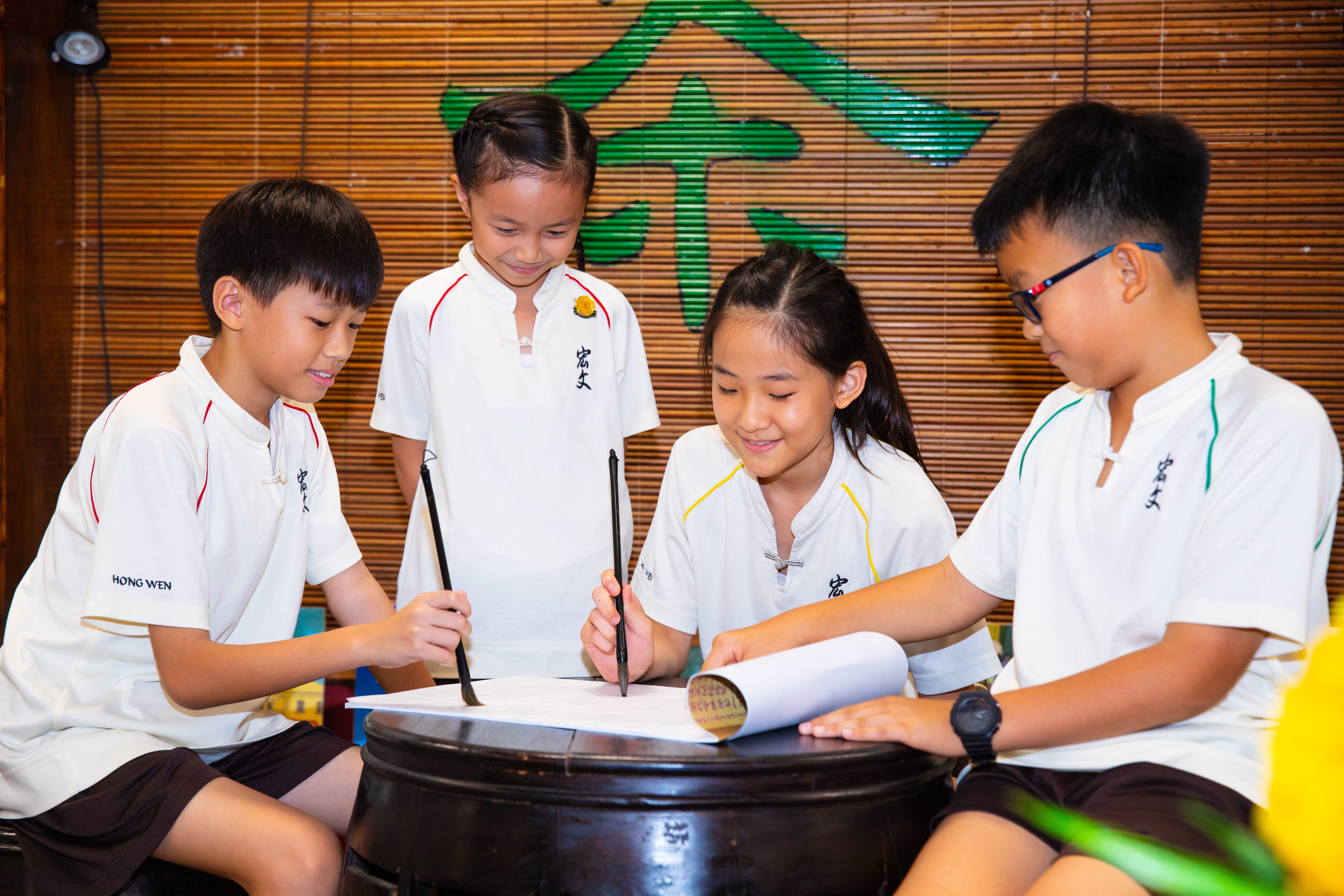 Four children in white uniforms use brushes to write on paper over a round table, with a bamboo wall behind.
