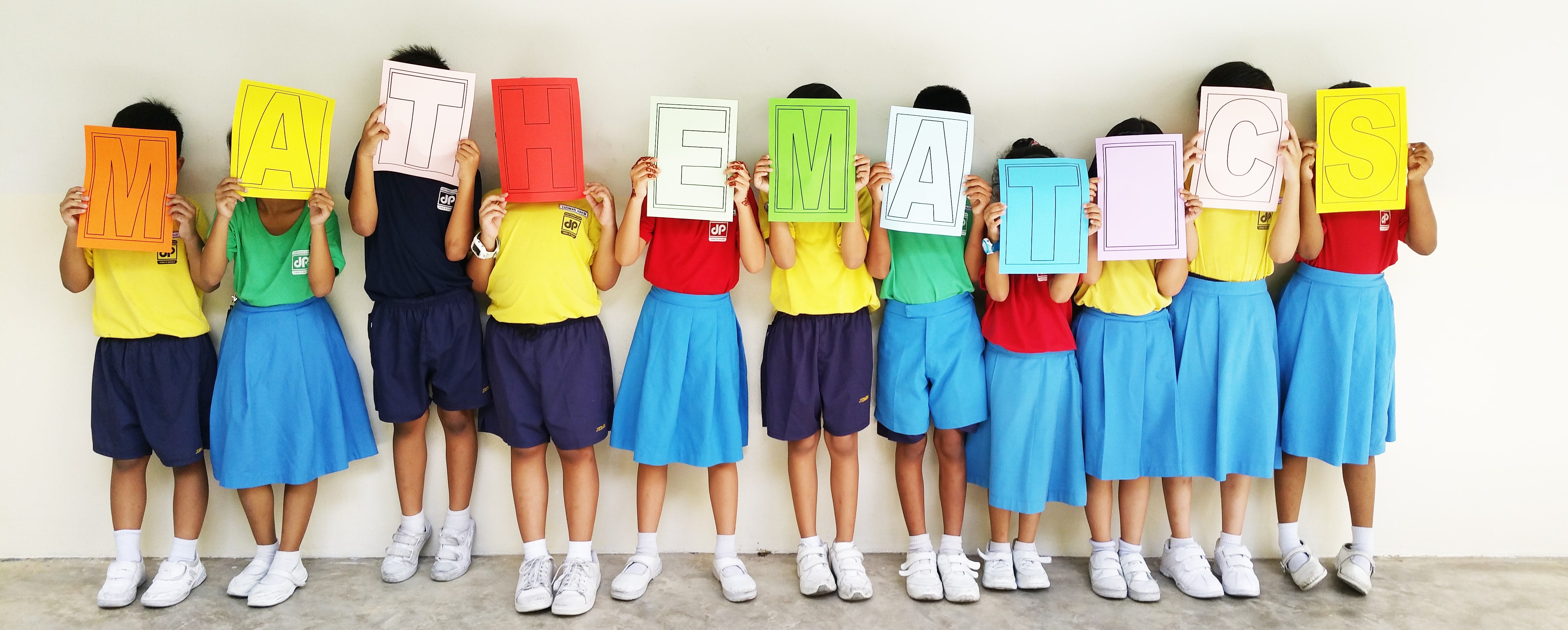A row of children in colorful uniforms each holding a letter card spelling MATHEMATICS over their faces.