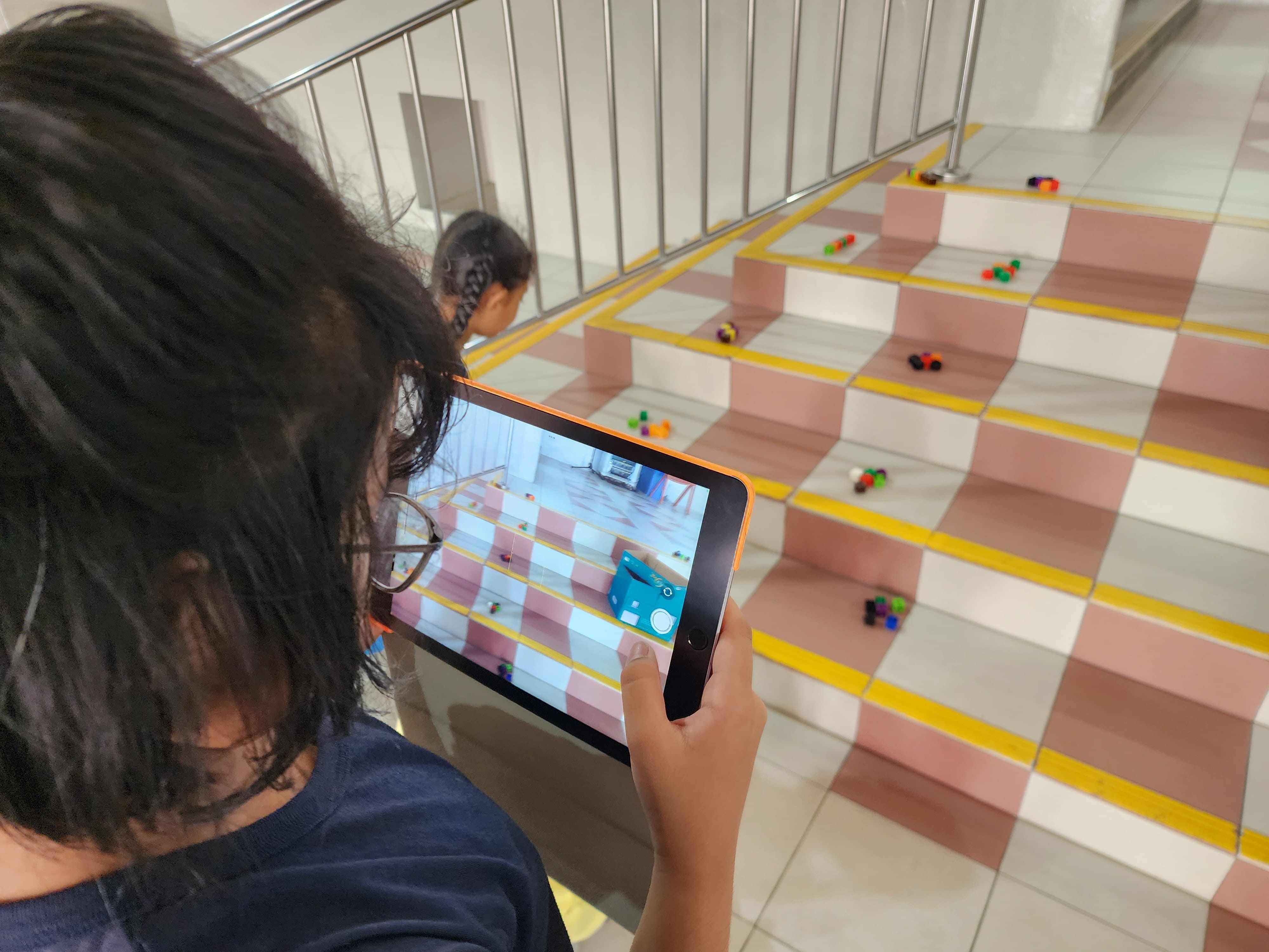 Child with glasses holds a tablet, photographing colorful toys scattered on pink and yellow tiled stairs.
