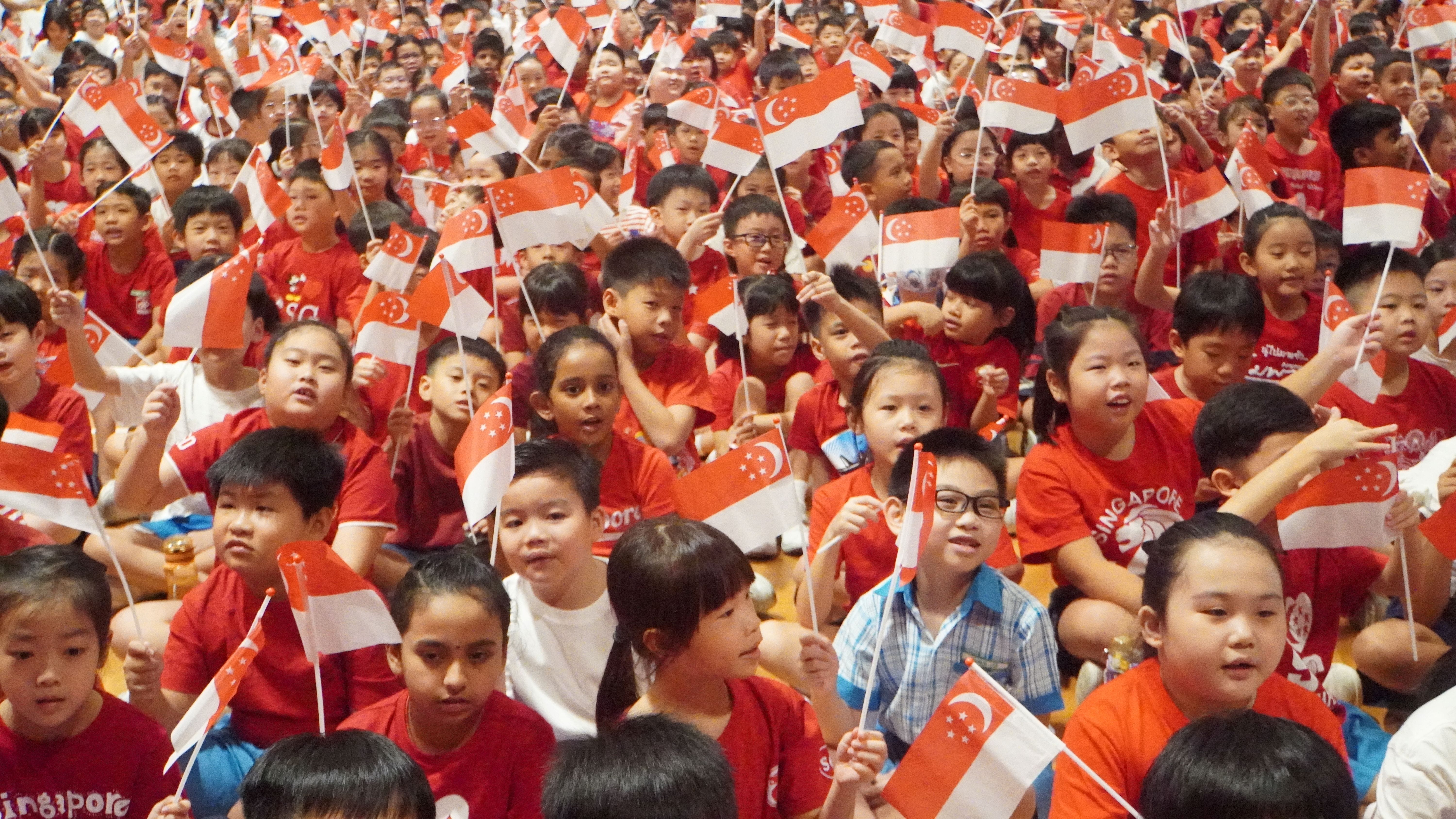 Students cheering and waving their mini flags proudly as they watch the National Day concert.