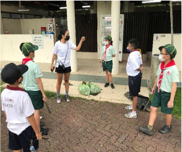 A woman in a mask instructs a group of masked children in scout uniforms outdoors near filled green bags.