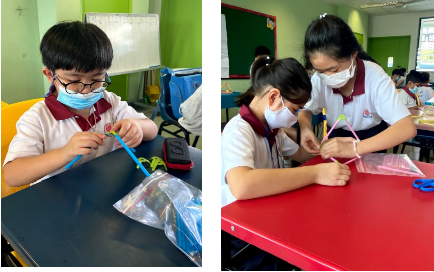 Students wearing masks work on a craft activity using colorful straws at classroom desks.