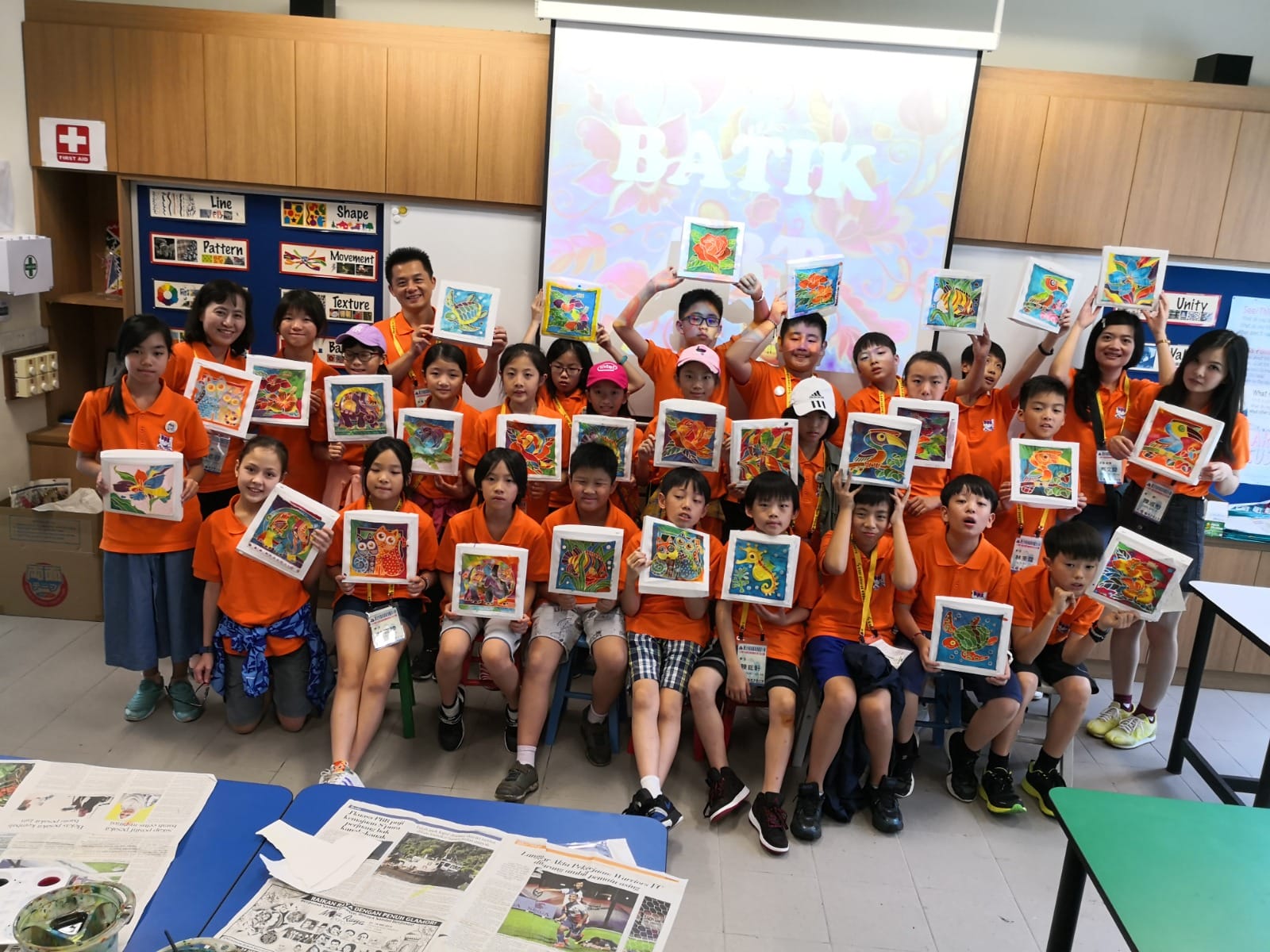 A group of children in orange shirts hold up colorful batik artwork in a classroom with a Batik-themed projector screen.