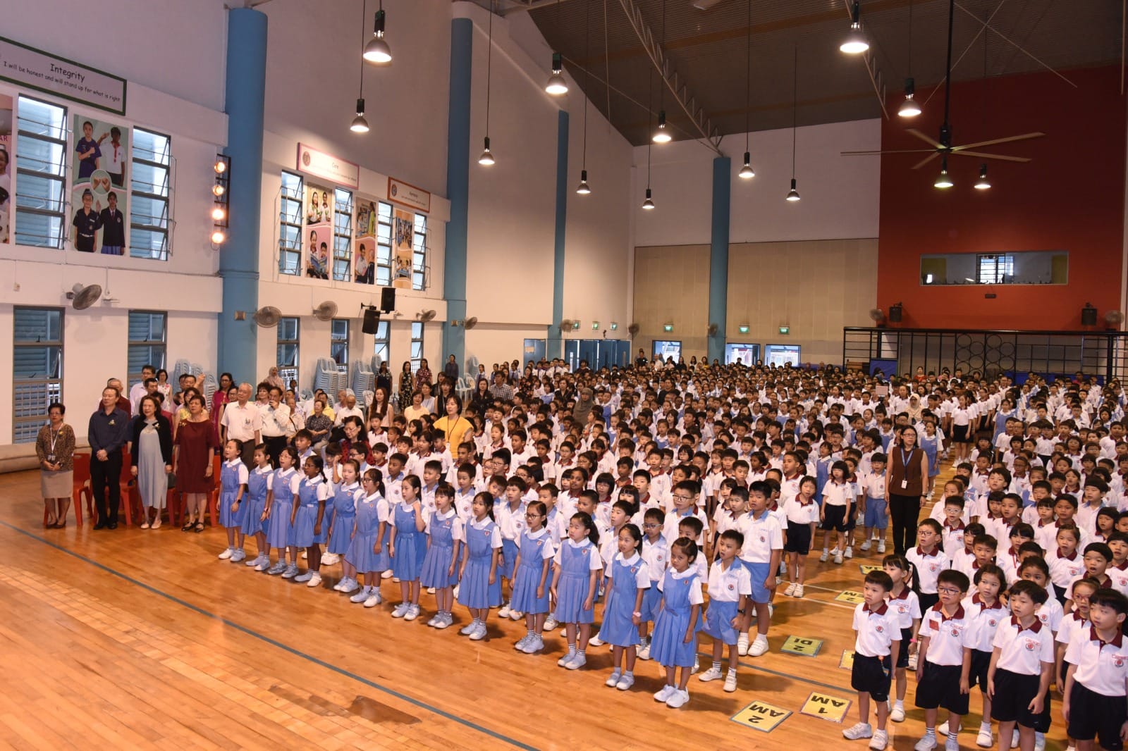 Hundreds of uniformed primary school students stand in rows in a large school hall during an assembly.