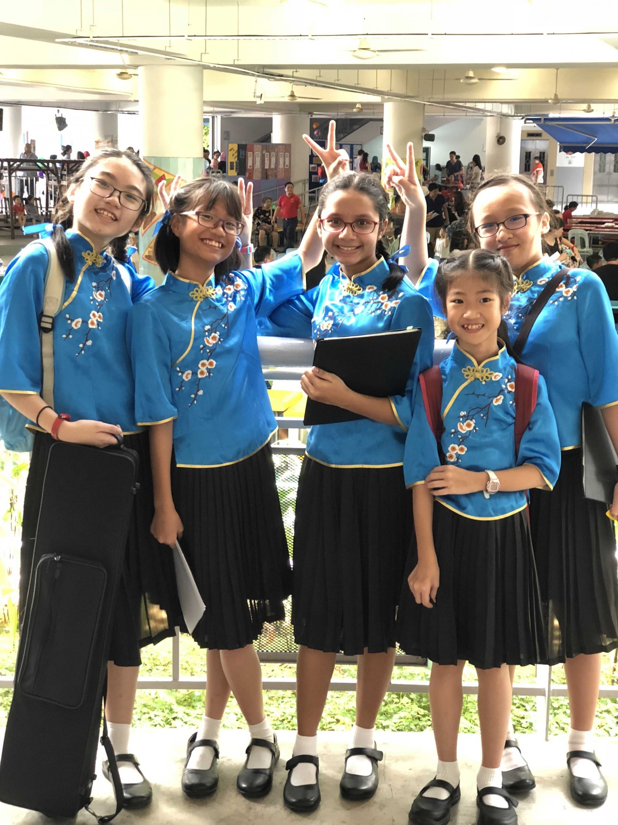 Five girls in matching blue floral uniforms and black skirts smile and pose with peace signs indoors.