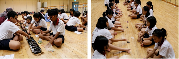 Children in white shirts sit cross-legged on a wooden floor, engaged in a hands-on activity with small objects.