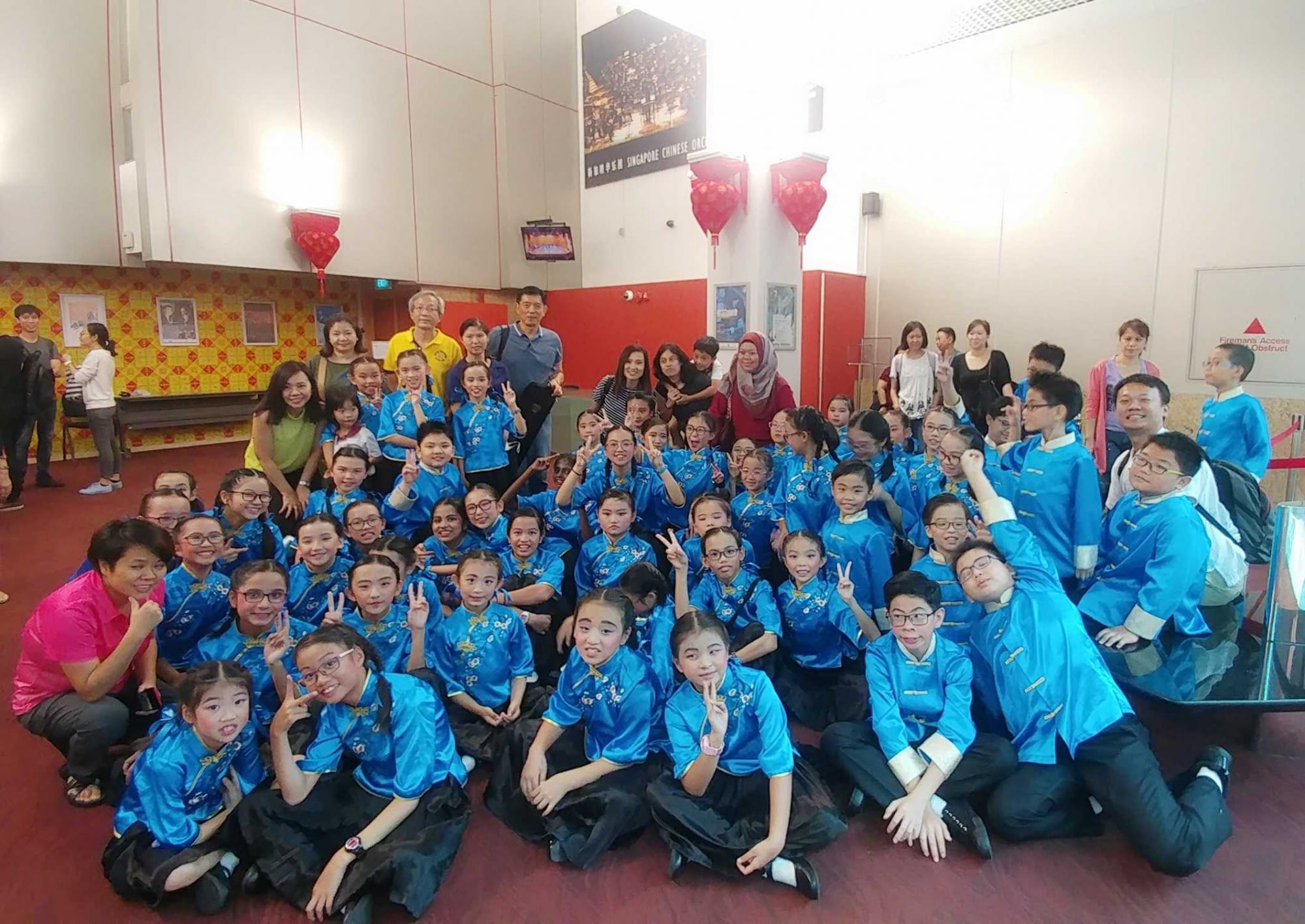 Large group of children in blue traditional Chinese outfits posing with peace signs indoors with red lanterns overhead.