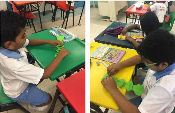 Two school boys in white uniforms fold green paper at colorful classroom desks.