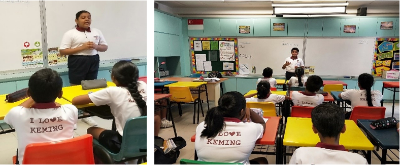 A student stands presenting to classmates in a colorful classroom, all wearing white shirts reading "I Love Keming."